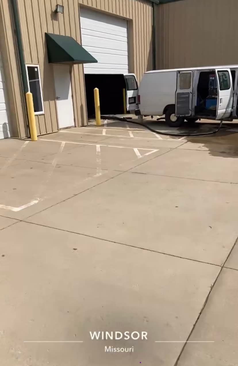 White van parked near a warehouse, with hose extending inside. Concrete area has parking space markings. Windsor, Missouri.