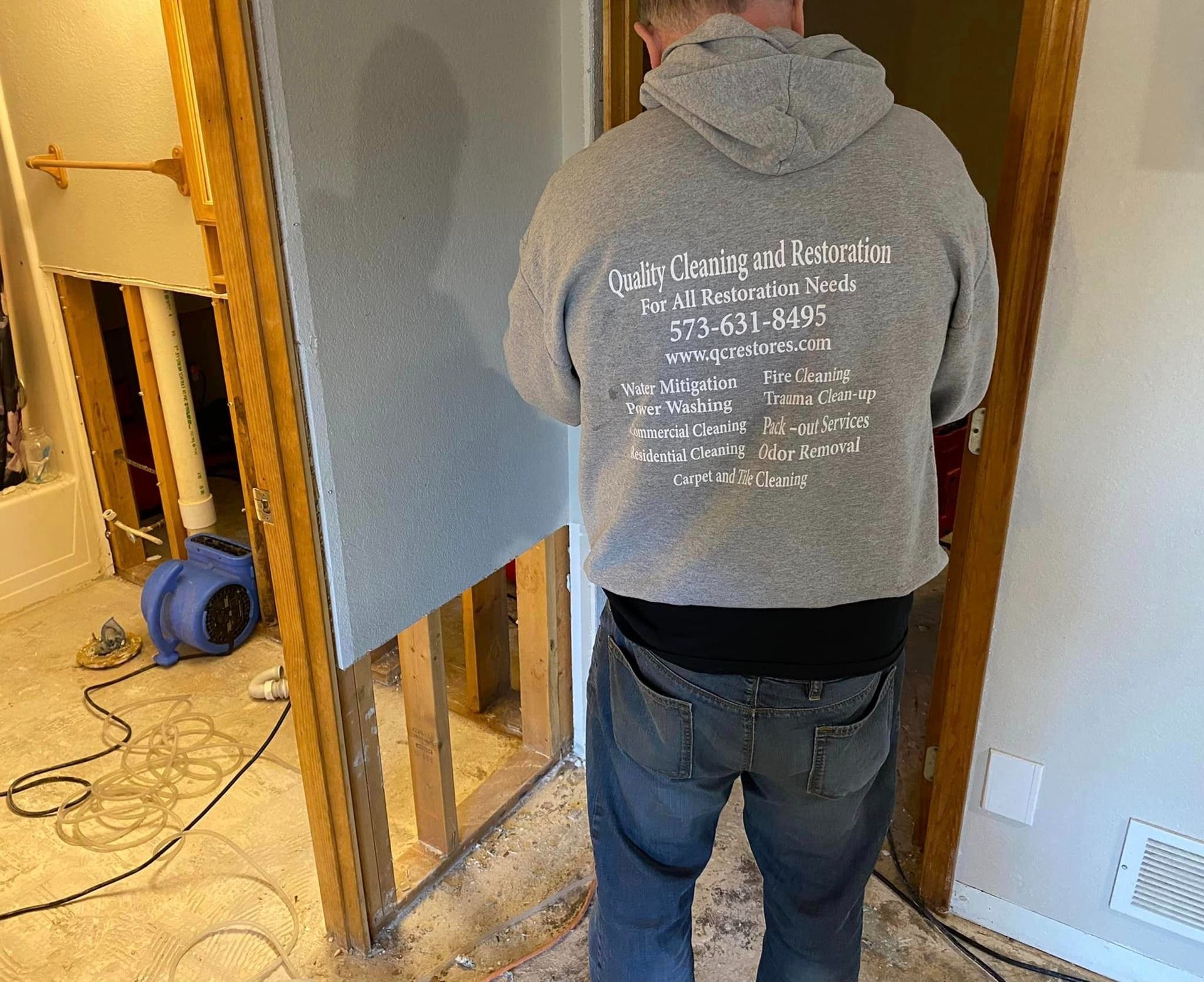 Man installing drywall in a room, wearing a gray hoodie, standing near an open doorway.