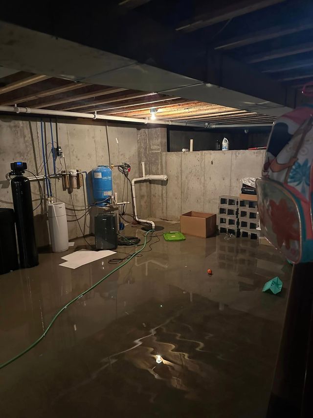 Flooded basement. Water covers floor, machinery and storage visible, concrete walls and exposed ceiling.