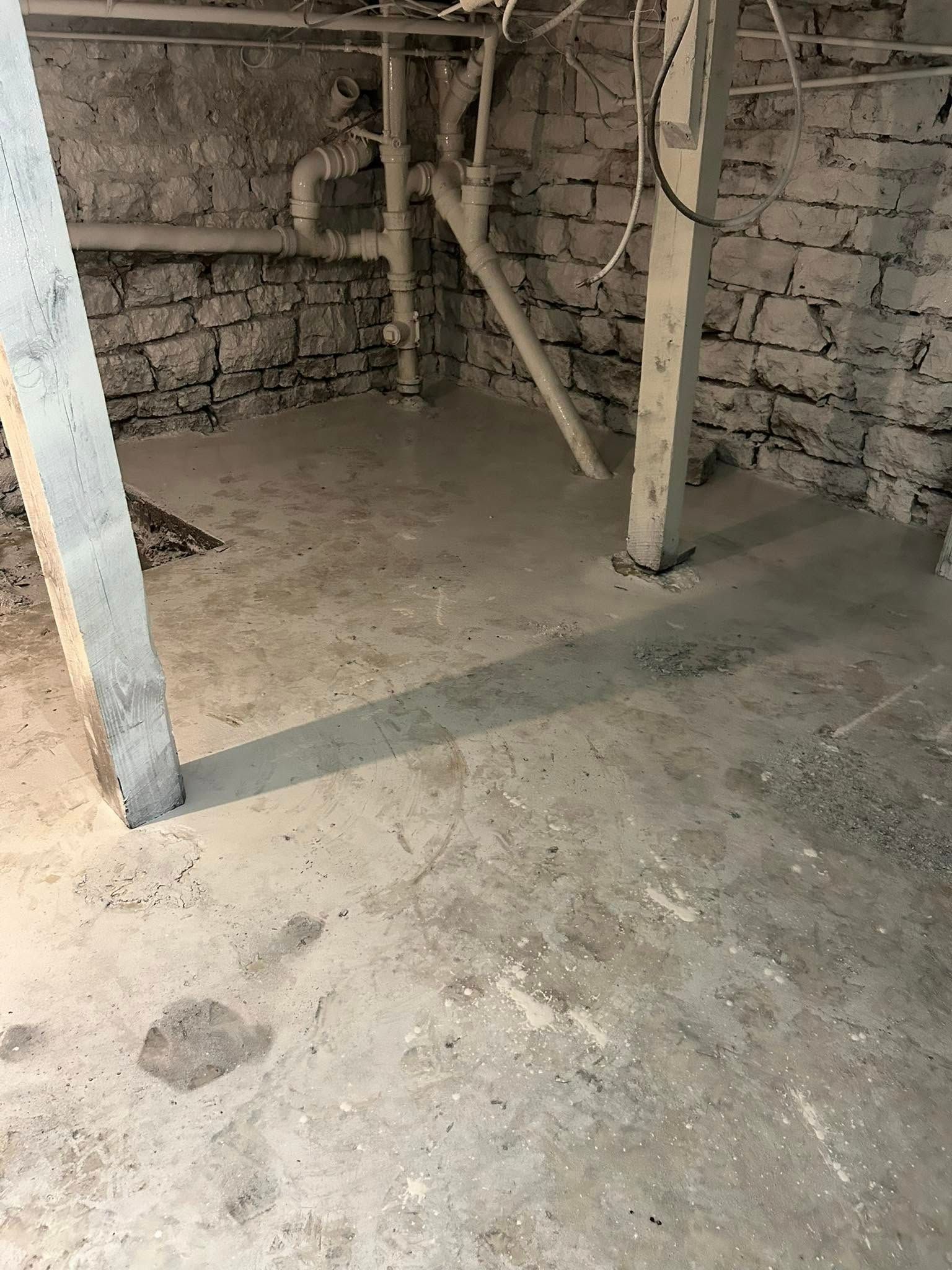 Concrete basement with exposed brick walls, plumbing, and support beams.