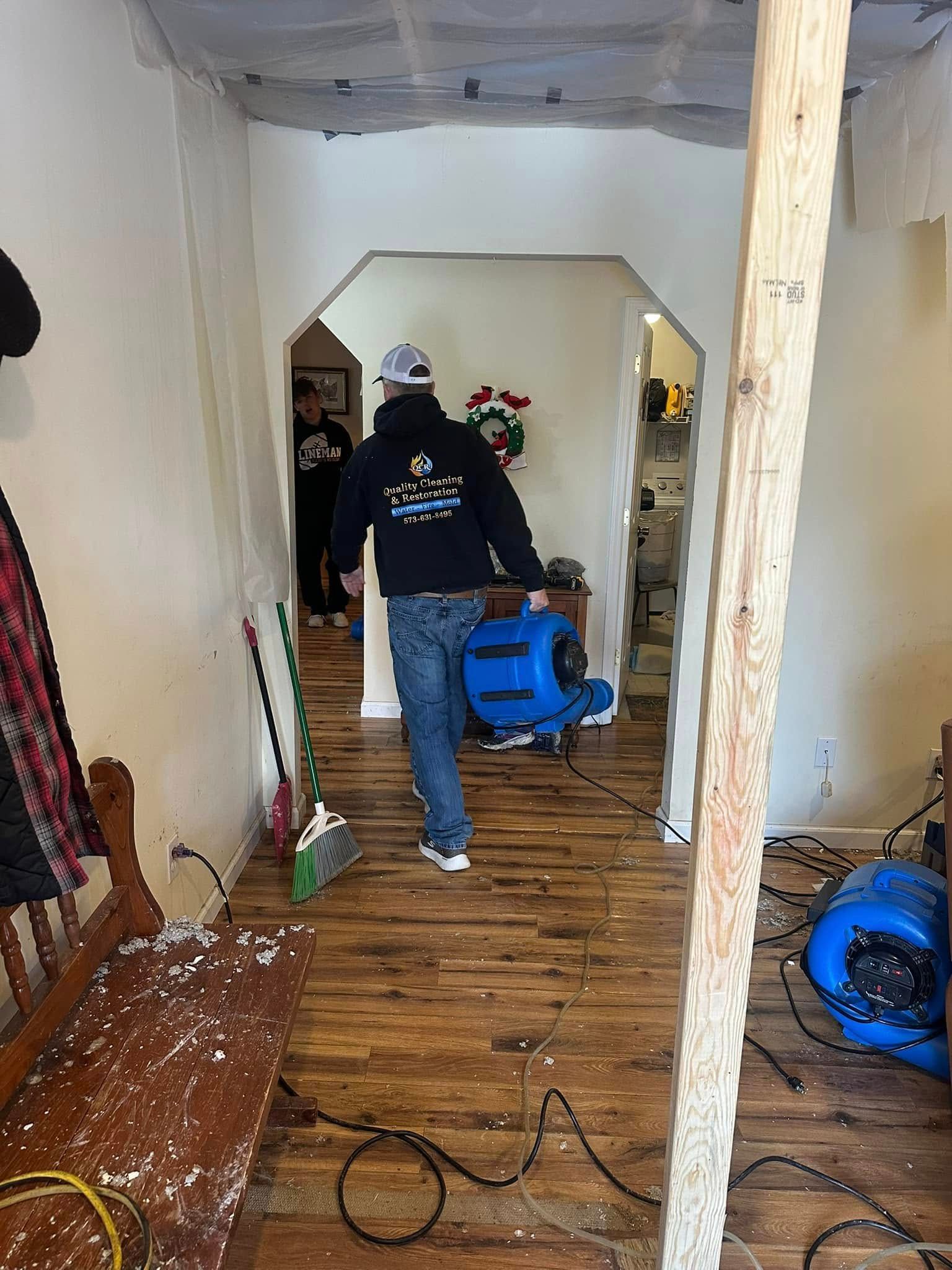 Person carries a blue air mover through a room with water damage. Brown floors and white walls.