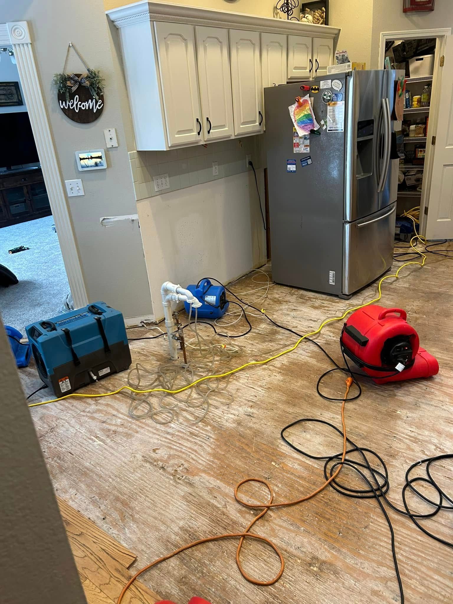 Kitchen with flood damage, drying equipment deployed on wet floor near cabinets, refrigerator, and door.