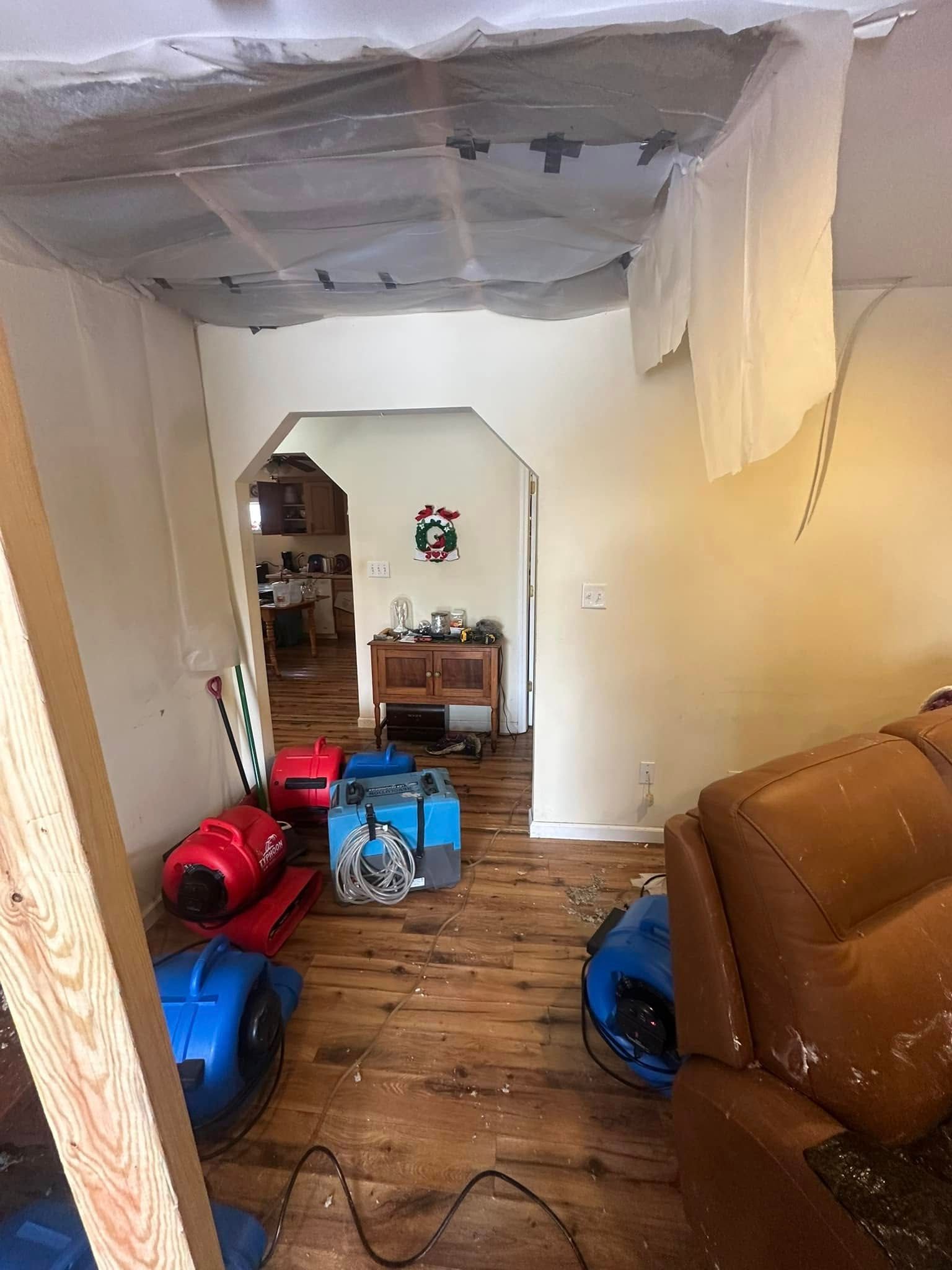 Debris-covered room with water damage; drying equipment on floor, doorway leads to another room.