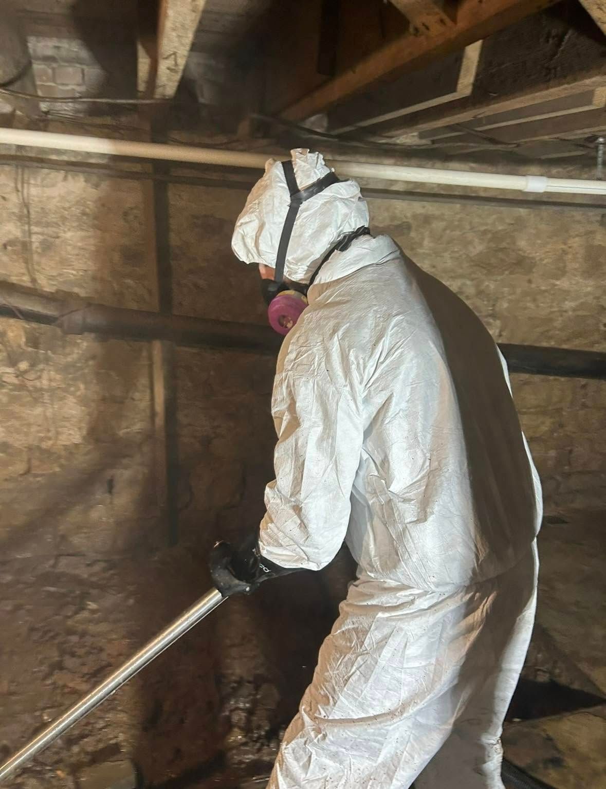 Person in hazmat suit sprays in a crawl space with brick walls.