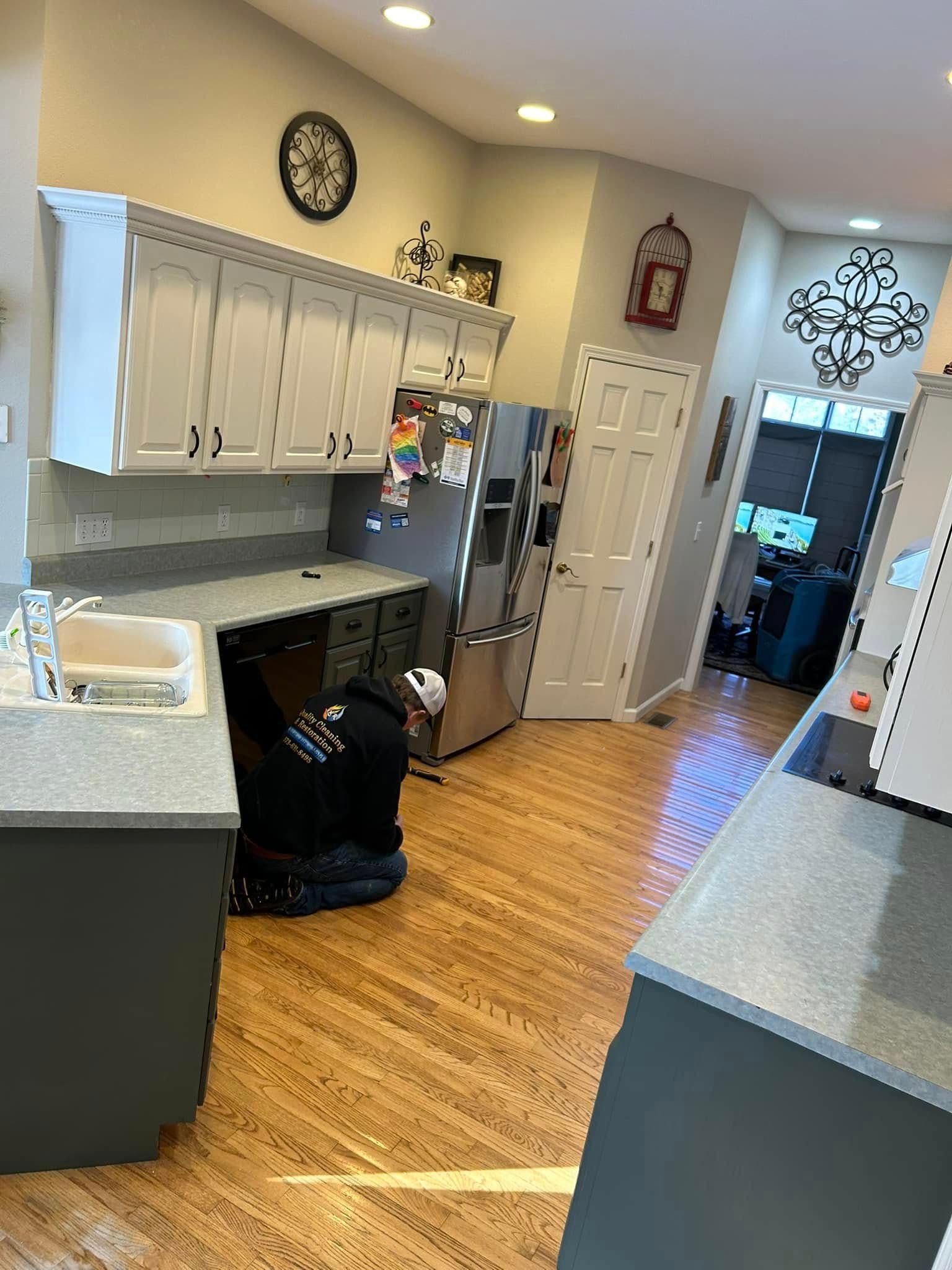 Kitchen with grey cabinets, appliances, and a person working on the floor. Wood flooring.