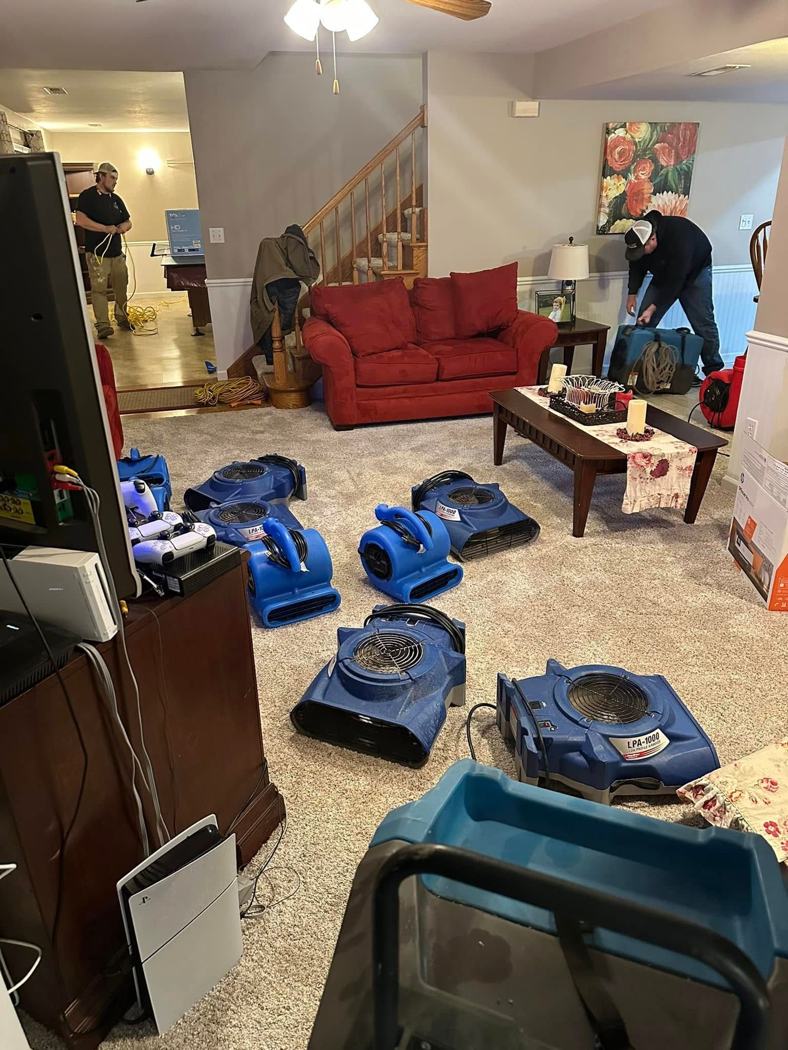 Flood damage in a living room; several blue air movers on the wet floor, two people assess damage near sofa and stairs.