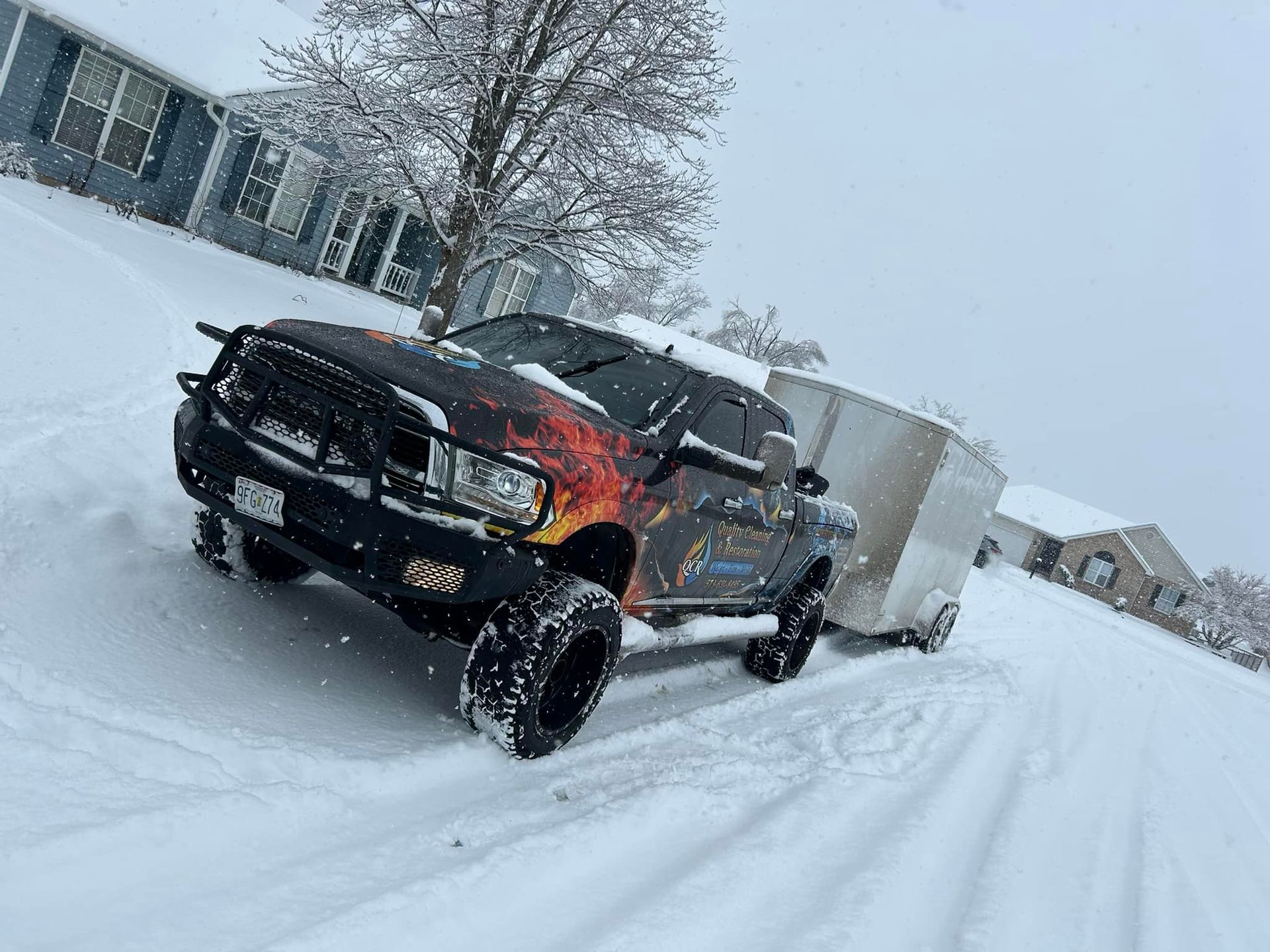 Black truck pulling a trailer in snowy conditions, parked on a snowy hill near houses.
