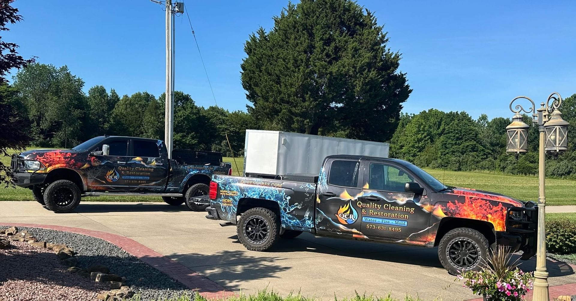 Two customized trucks parked in a driveway; one black and one silver, both with colorful graphic wraps.