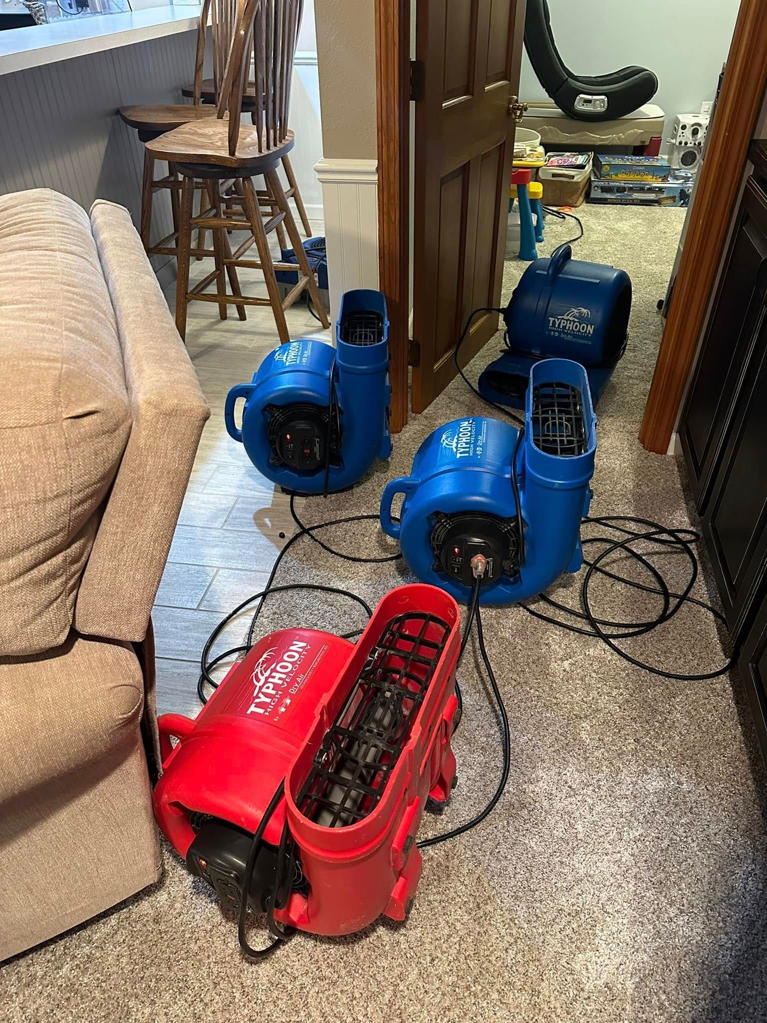 Blue and red air movers set up in a room with a sofa, chair, and open doorway; presumably drying out the space.