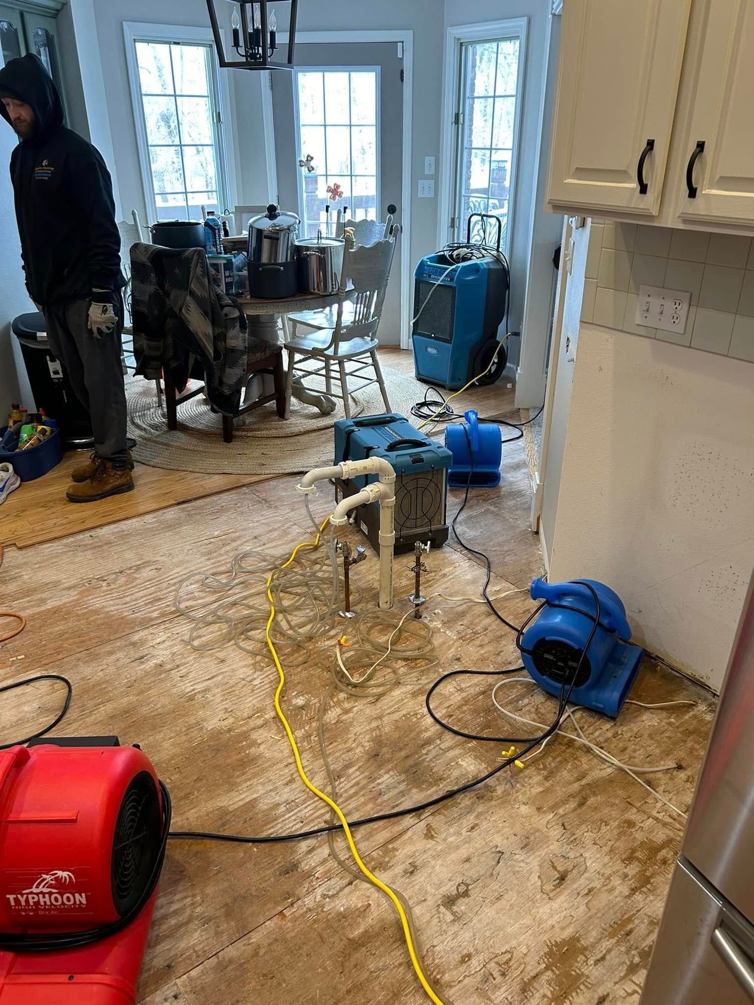 Man in flooded kitchen with drying equipment: fans, dehumidifiers, hoses. Brown flooring is wet.