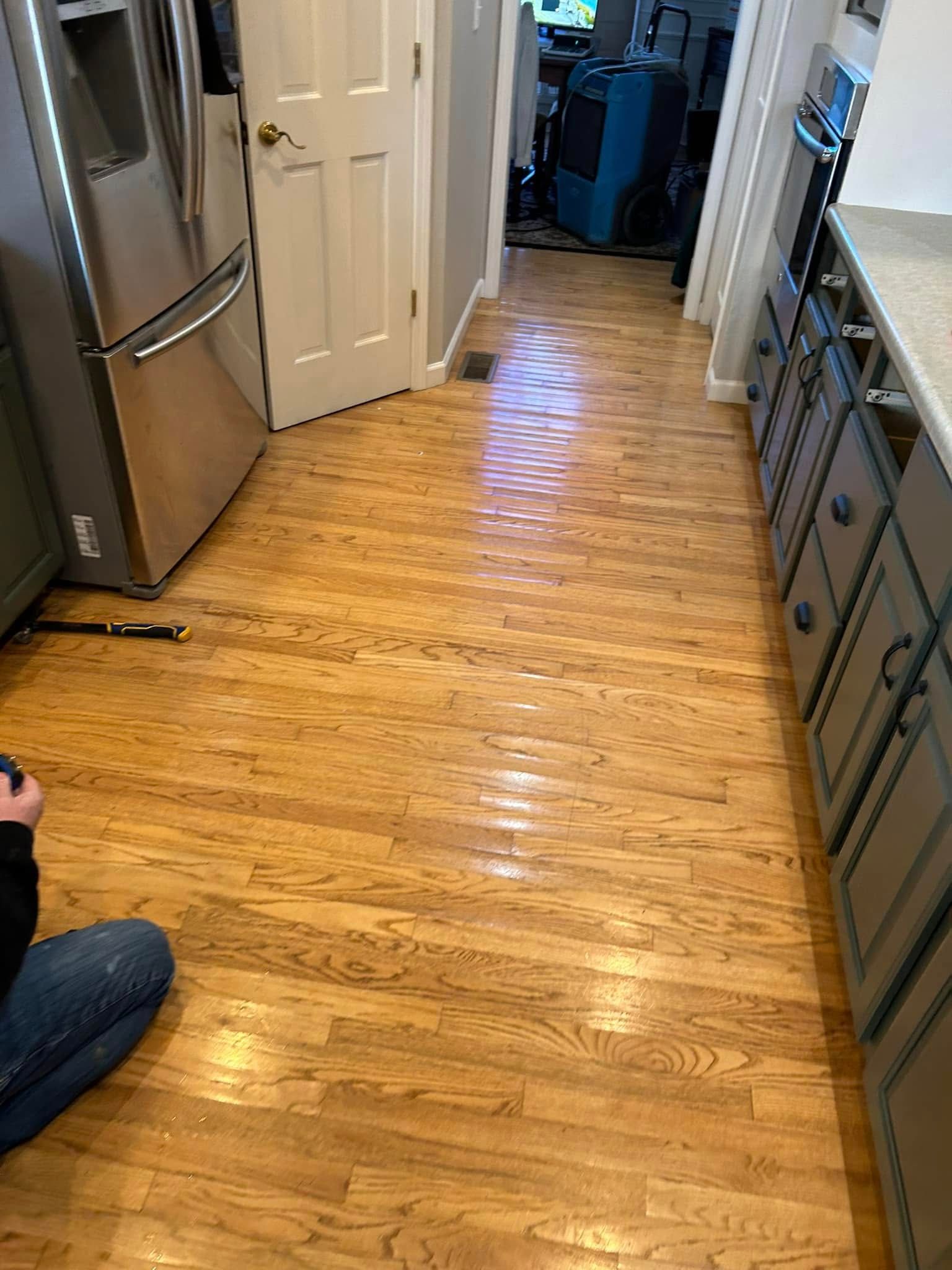 Wooden floor in kitchen with cabinets and appliances; person kneeling on floor.