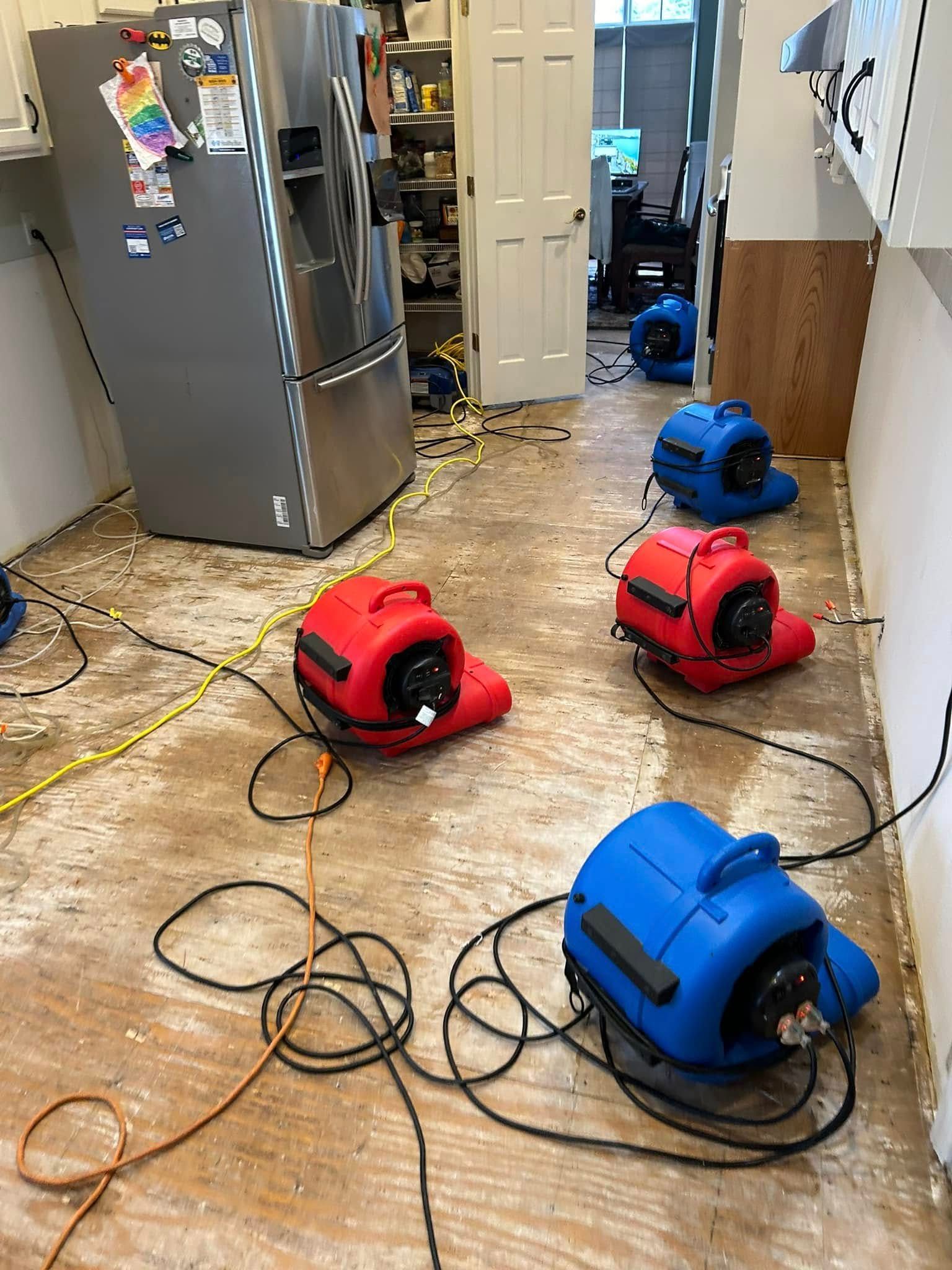 Red and blue air movers drying a kitchen floor after a water incident, near a refrigerator and open door.