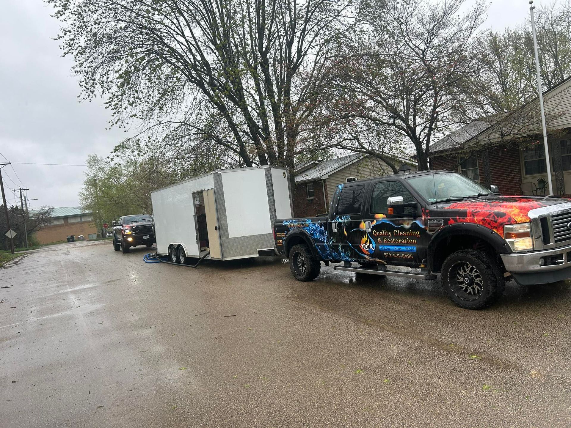 Truck towing a white enclosed trailer on a wet street. Other vehicle in distance, overcast sky.