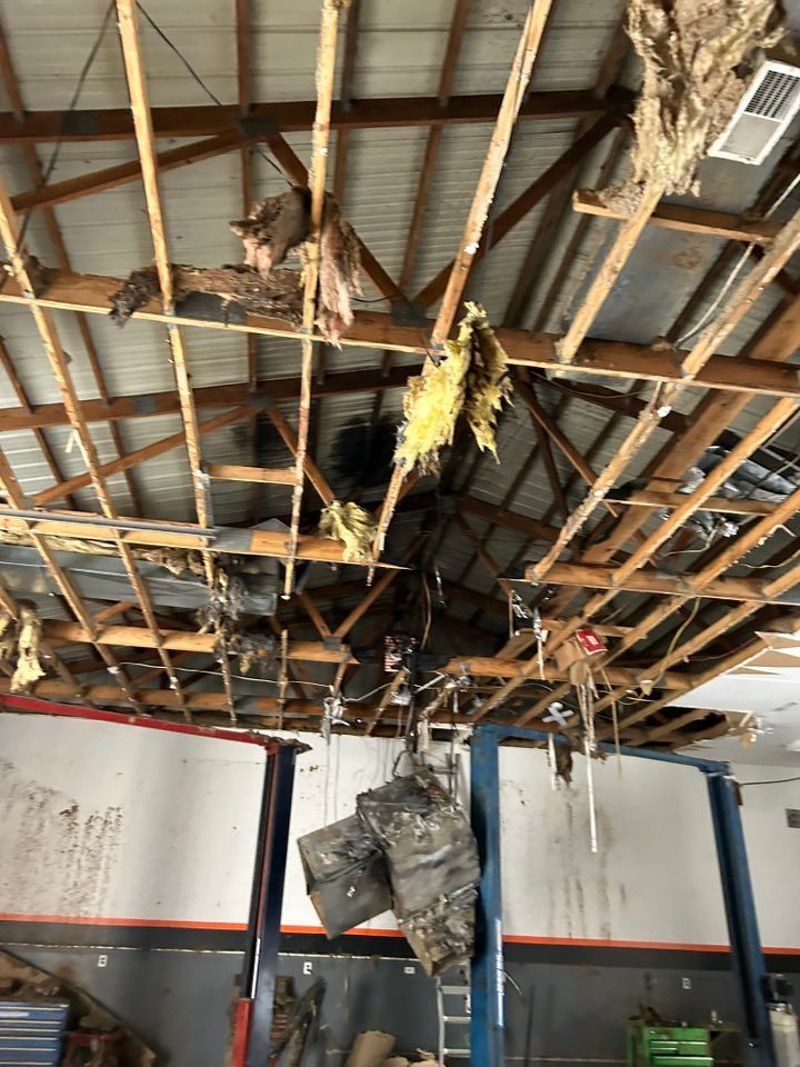 Damaged workshop ceiling with exposed beams, insulation, and debris. A car lift is visible below.