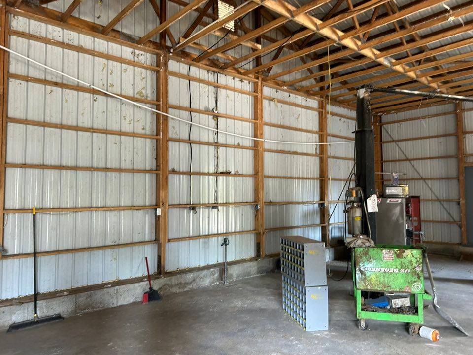 Interior of a barn with metal walls, wooden beams. A green machine, gray boxes, and a black pipe are visible.