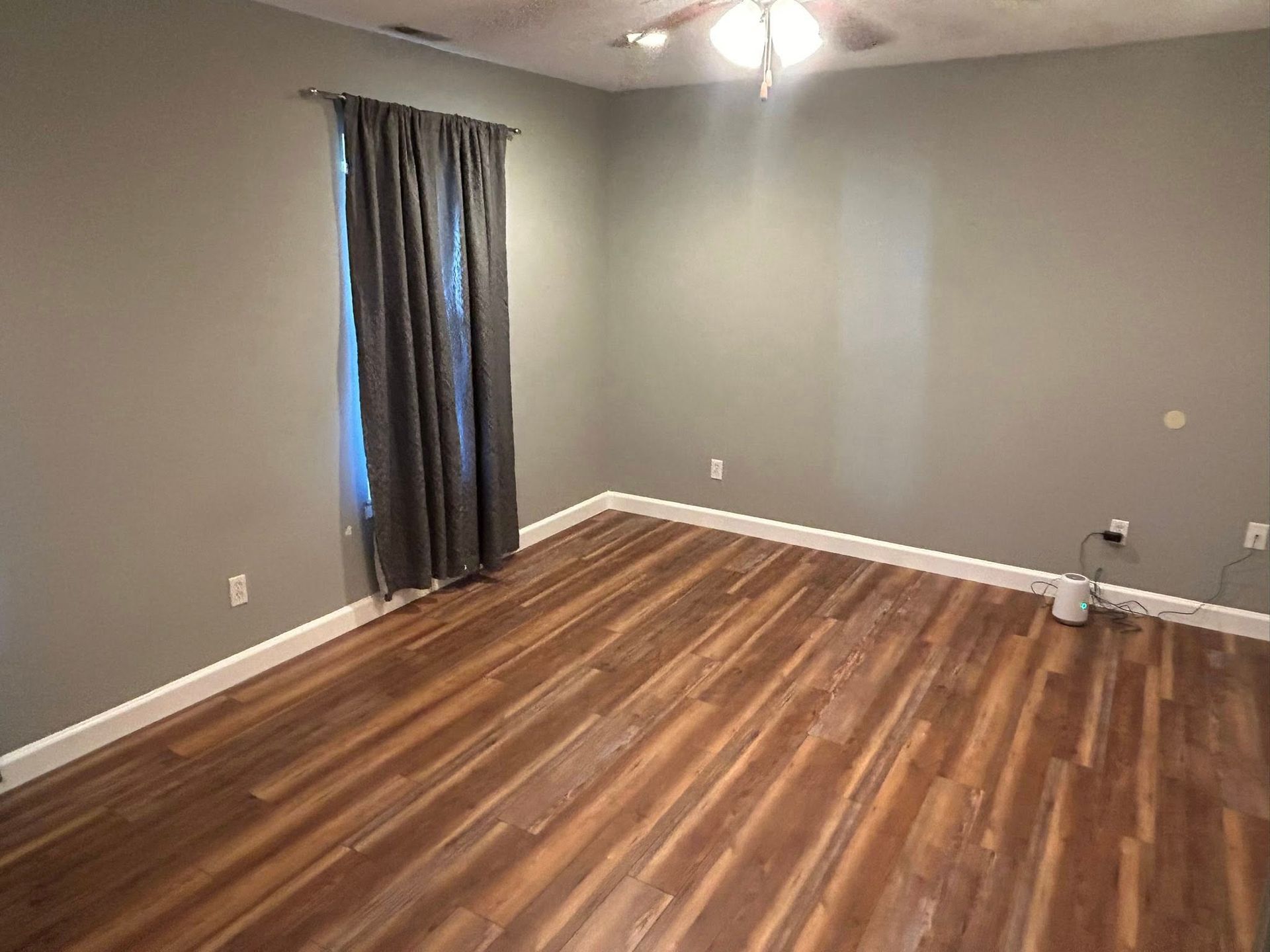Empty room with gray walls, hardwood floor, window with dark curtain, and ceiling fan.