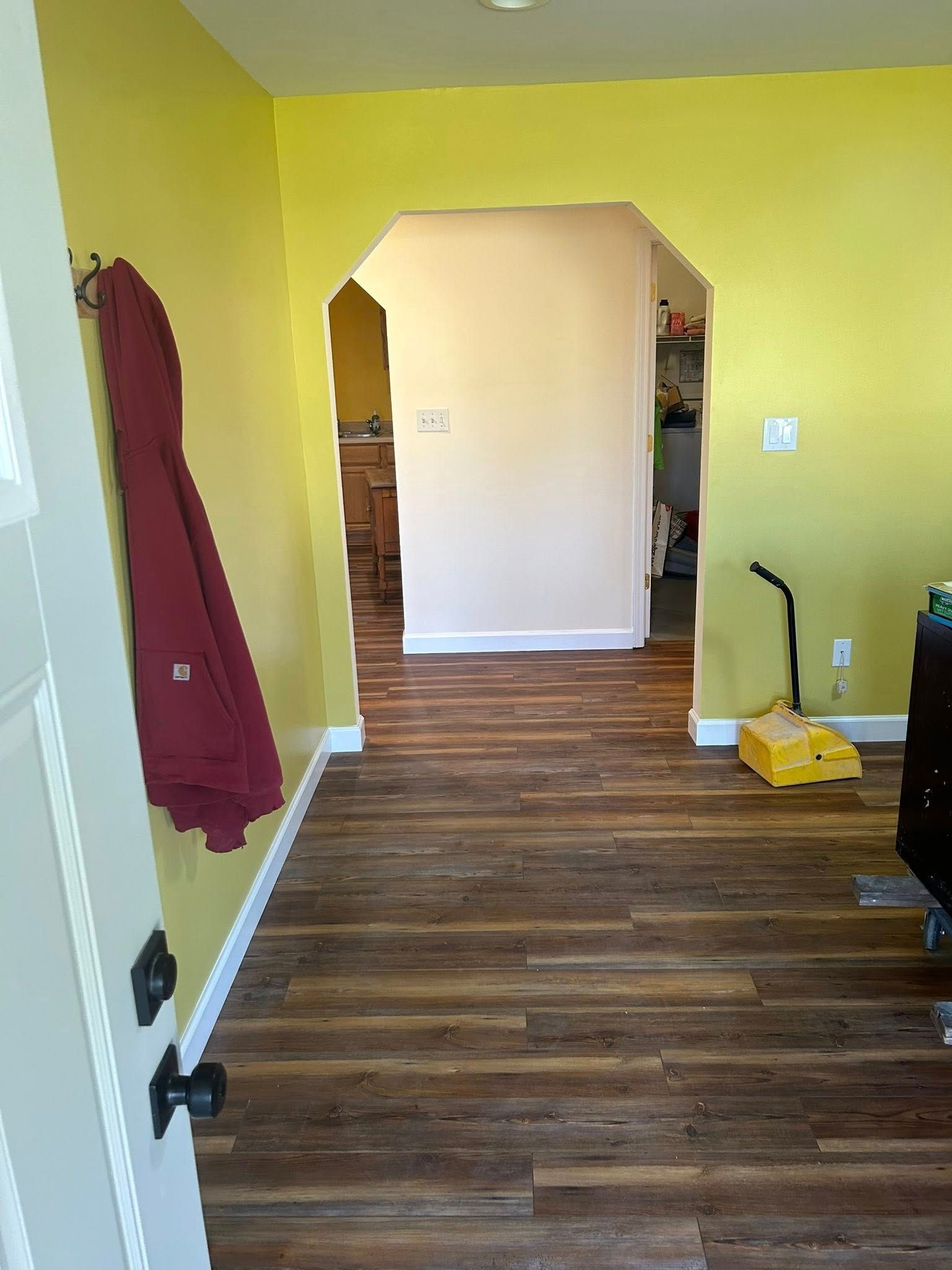 Hallway with yellow walls, wood-look flooring, arched doorway leading to another room, coat hanging on wall.