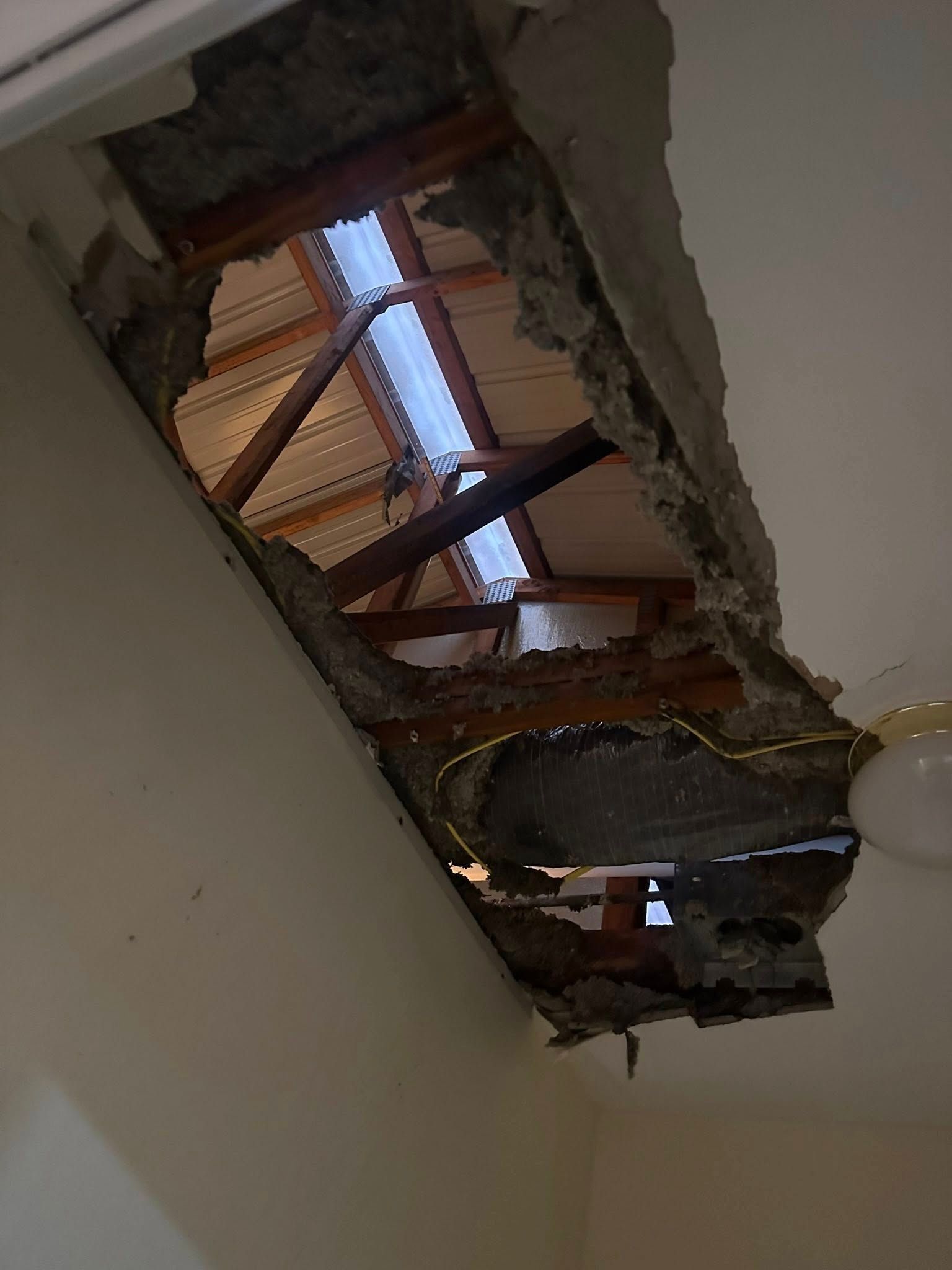 Hole in a ceiling, revealing roof structure and skylight. Gray concrete, wooden beams, bright light visible.