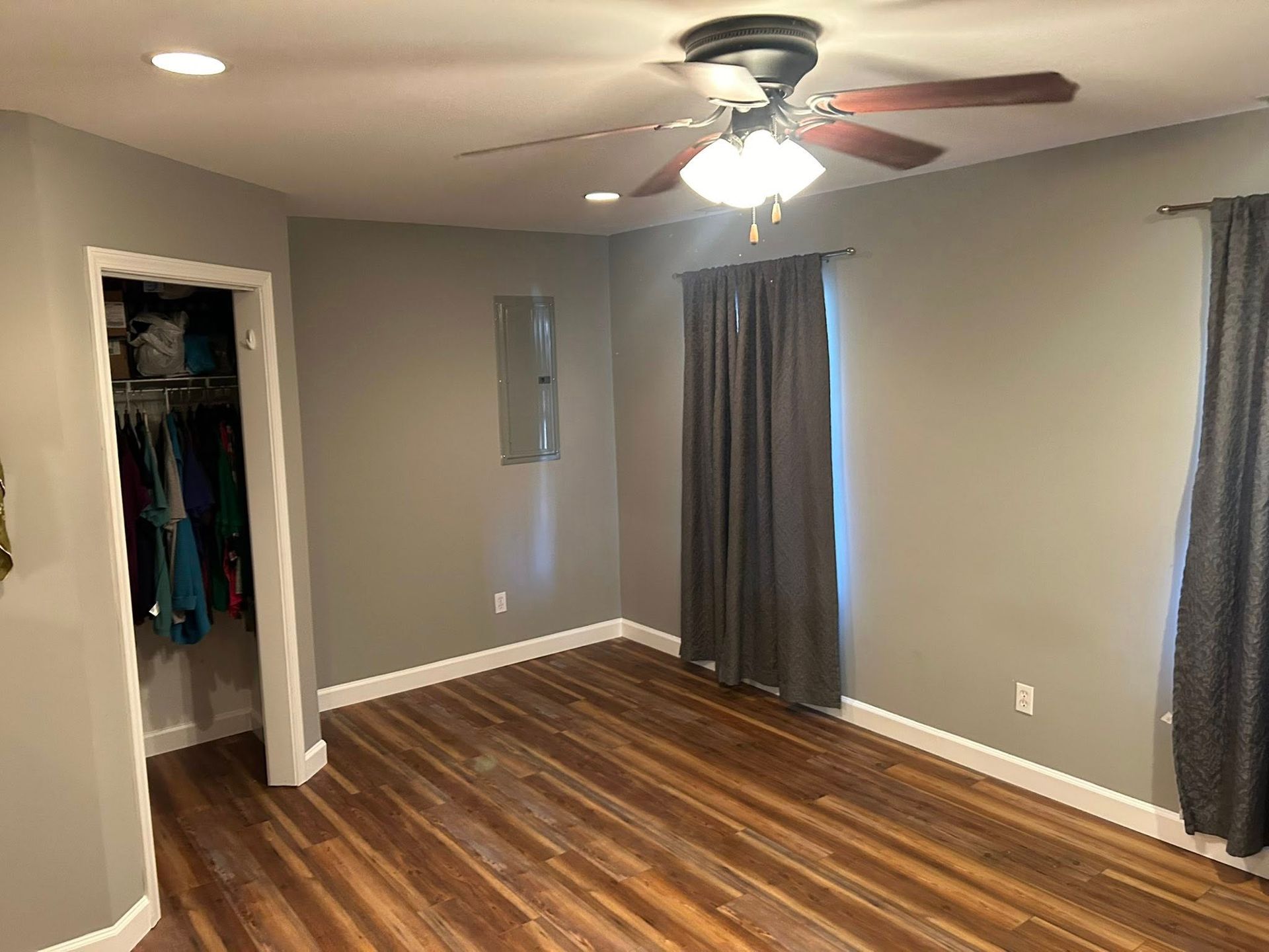 Bedroom interior with gray walls, wood-look floor, walk-in closet, and dark curtains. Ceiling fan installed.