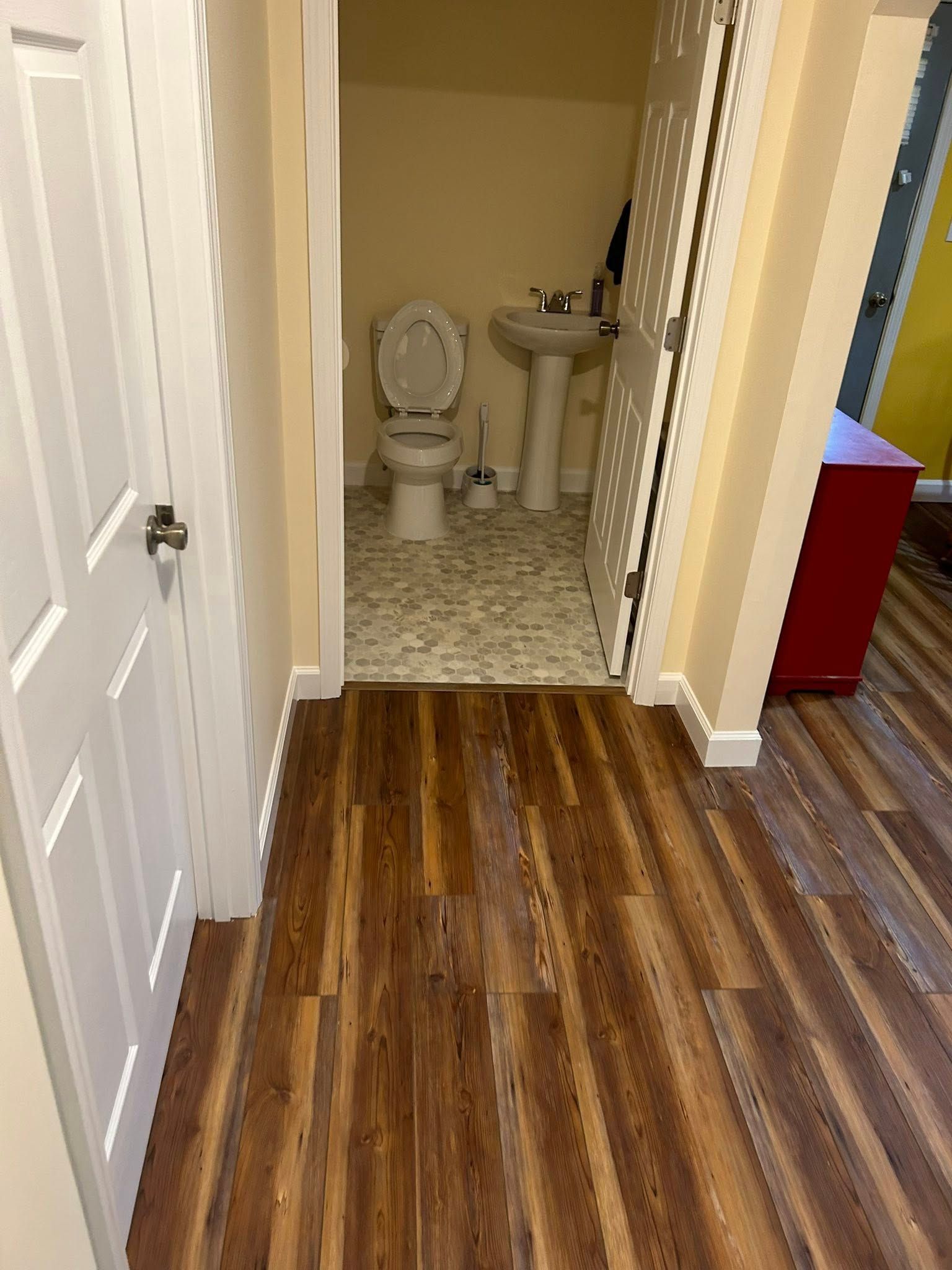Hallway with wood-look floor leading to bathroom with toilet and sink. White door on the left, open door on the right.