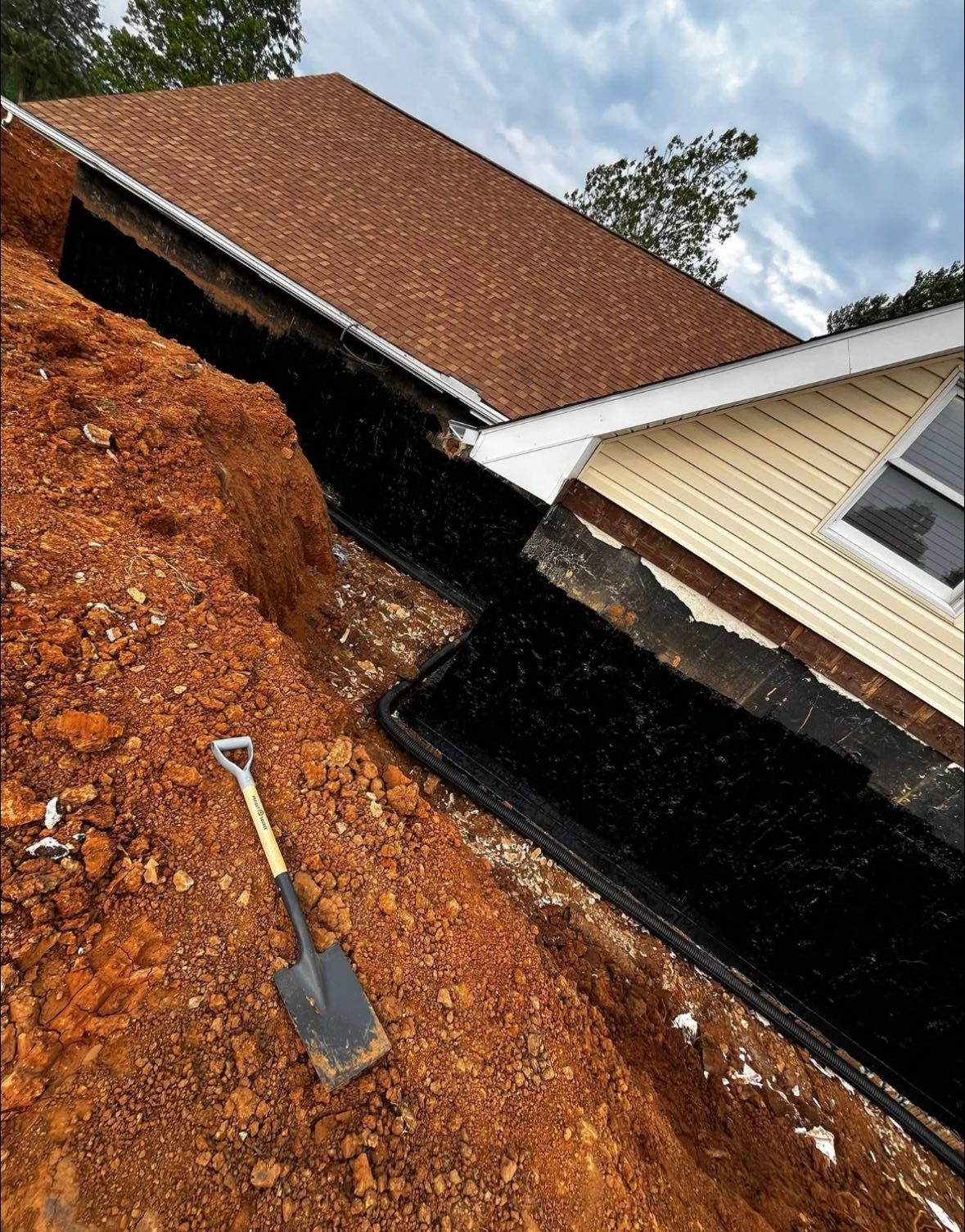 Trench dug along a building's foundation, exposing black waterproofing membrane. Shovel in the foreground.