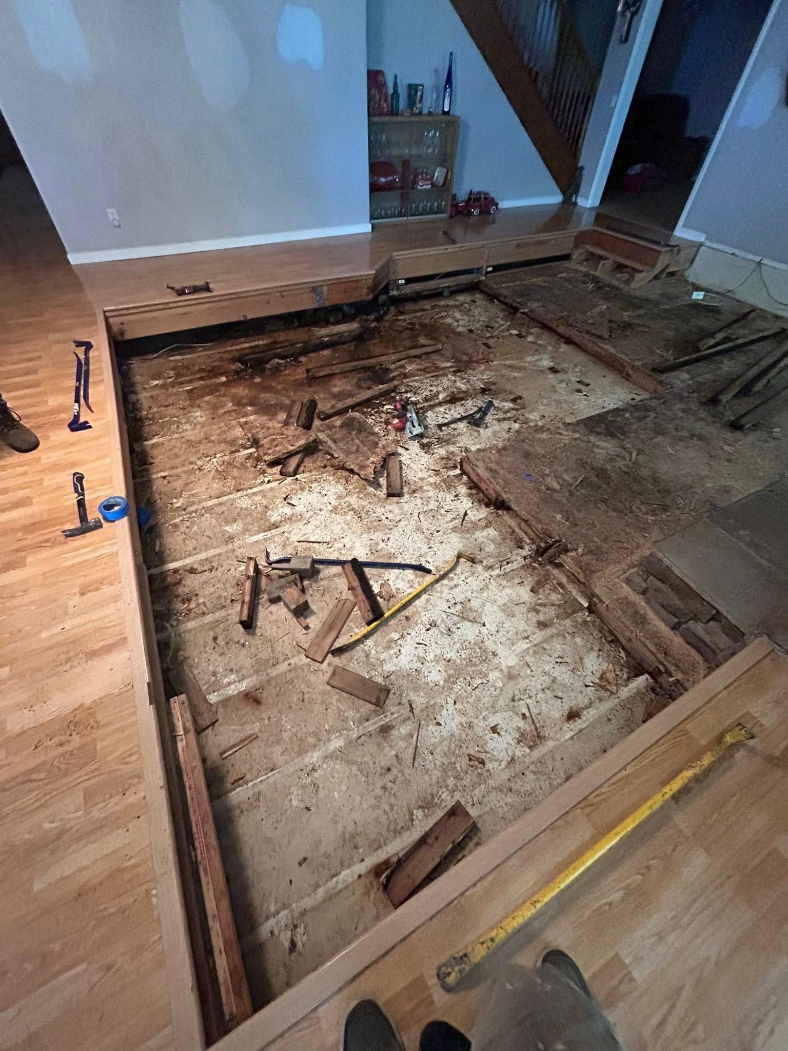 Floorboards removed, revealing subfloor. Tools and debris scattered across the exposed area.