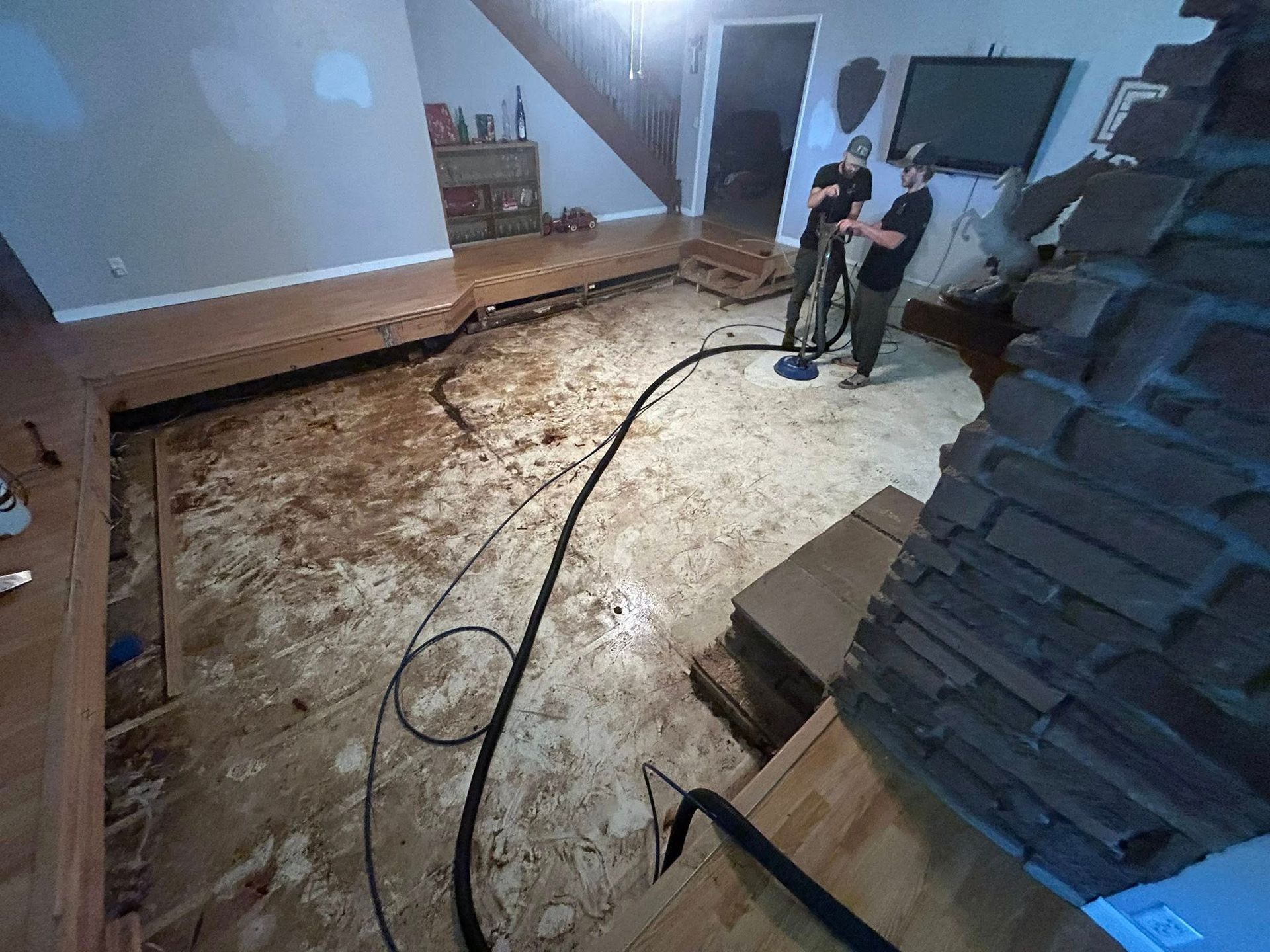 Men vacuuming debris from a flooded living room, damaged wood flooring, fireplace, stairs.