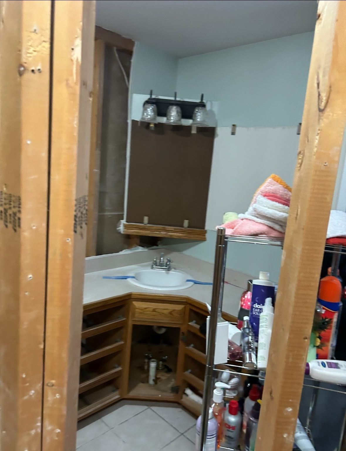 Bathroom under construction; view through wooden framing. Sink with cabinets, mirror, shelves with products.