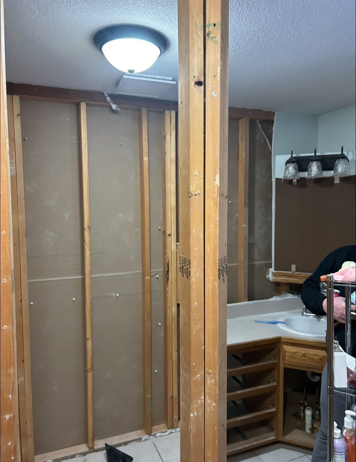 Bathroom interior during renovation, with exposed studs and partially removed walls. A person is visible.