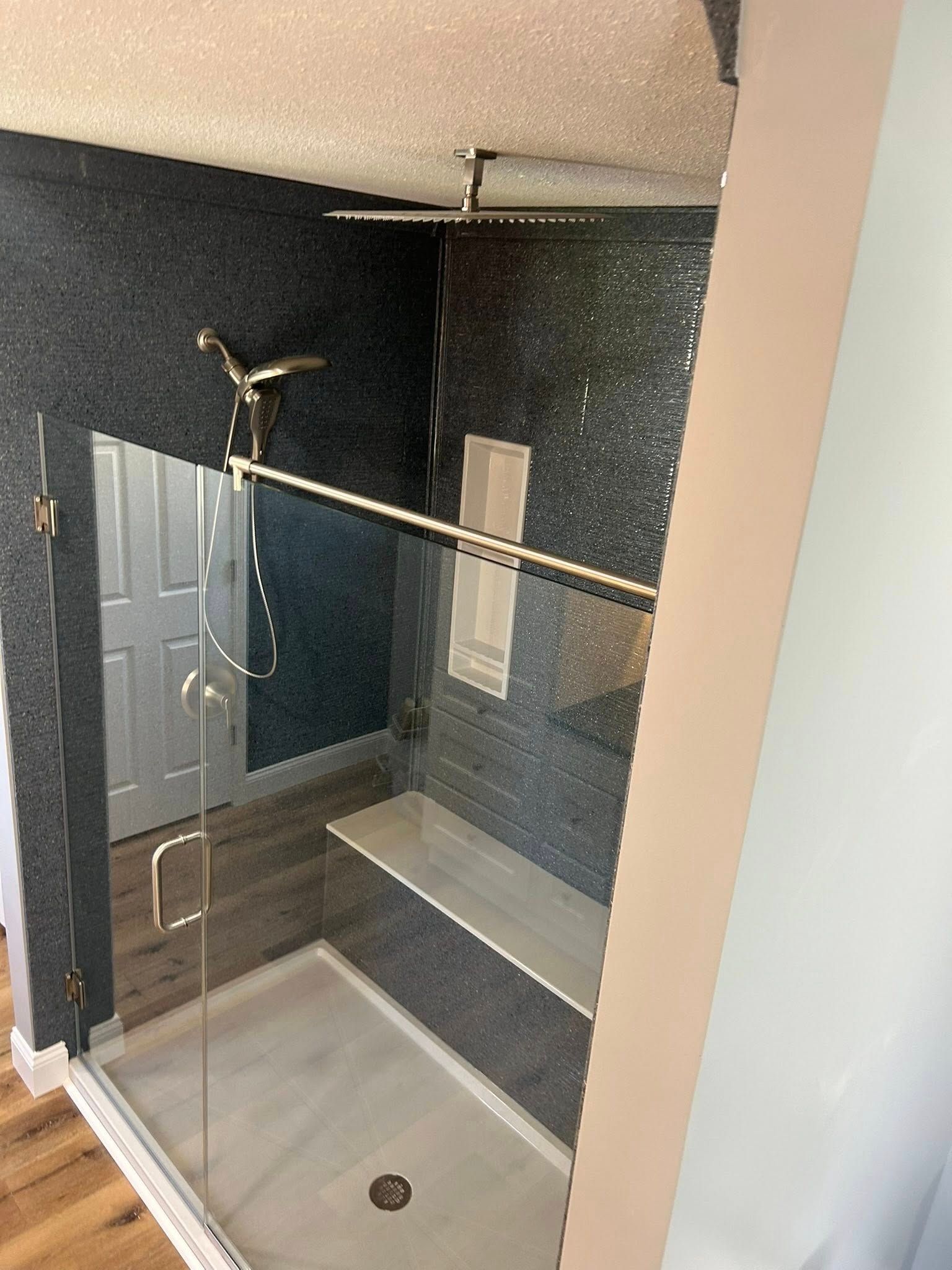 Modern shower with glass door, bench, and gray tile walls. Beige trim and light wood flooring.