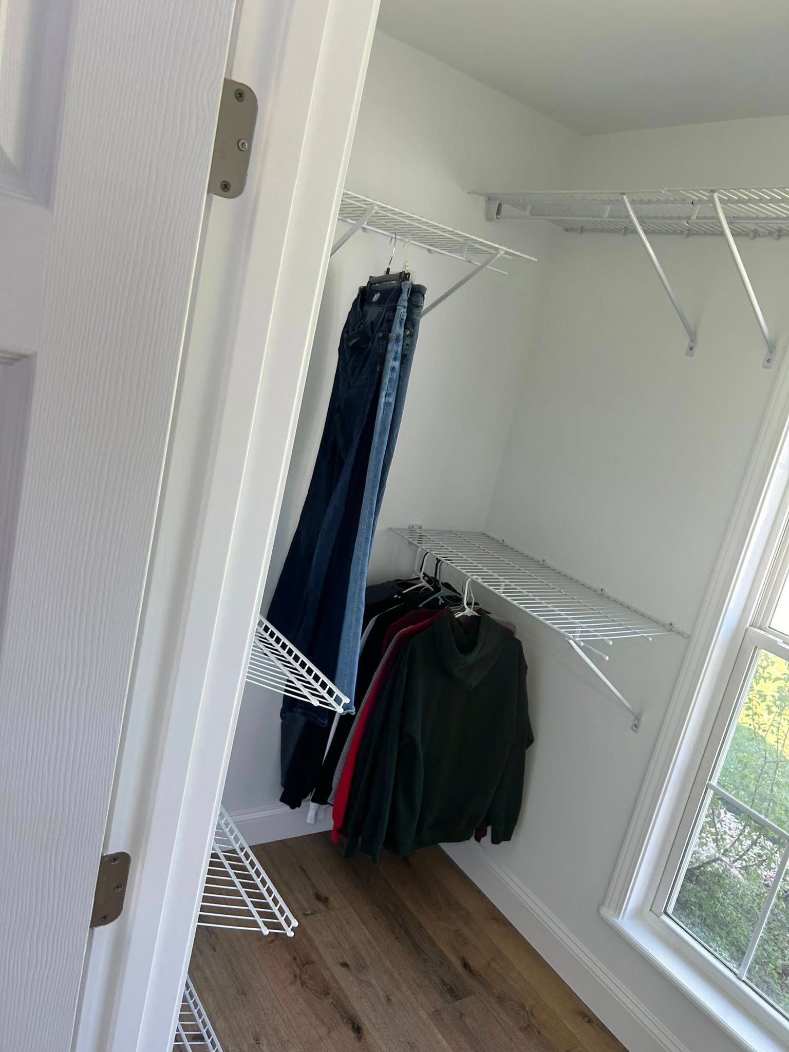 White closet with hanging clothes, wire shelving, wood floor, and a window.