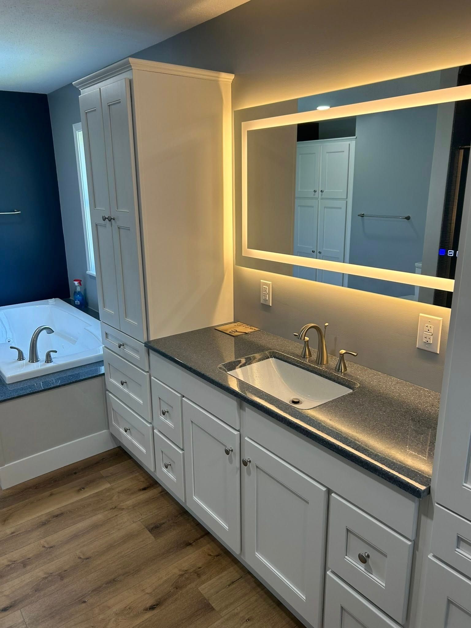 Bathroom with white cabinets, dark countertop, lit mirror, and a tub.