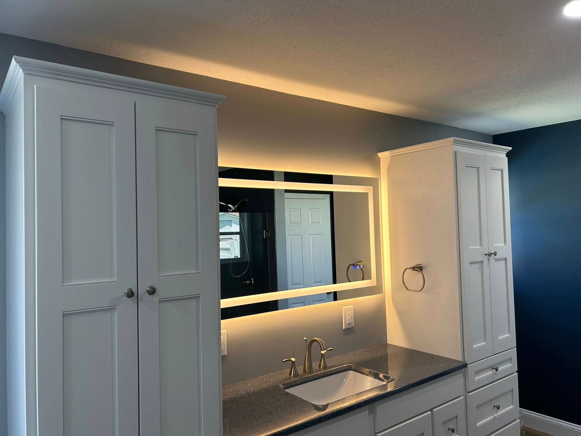 White bathroom vanity with built-in cabinets, a backlit mirror, and a granite countertop against a blue wall.