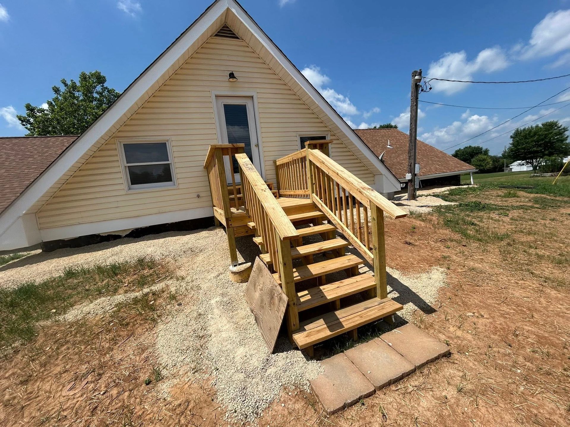 Wooden stairs leading up to the door of a beige A-frame house. Gravel landscaping. Sunny day.