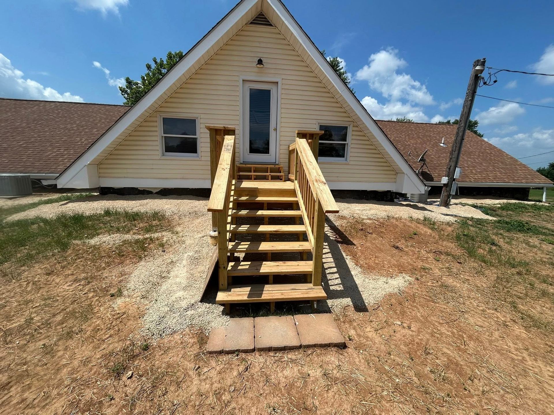 A-frame house with wooden stairs leading to a front door, set on a sloped yard with gravel.