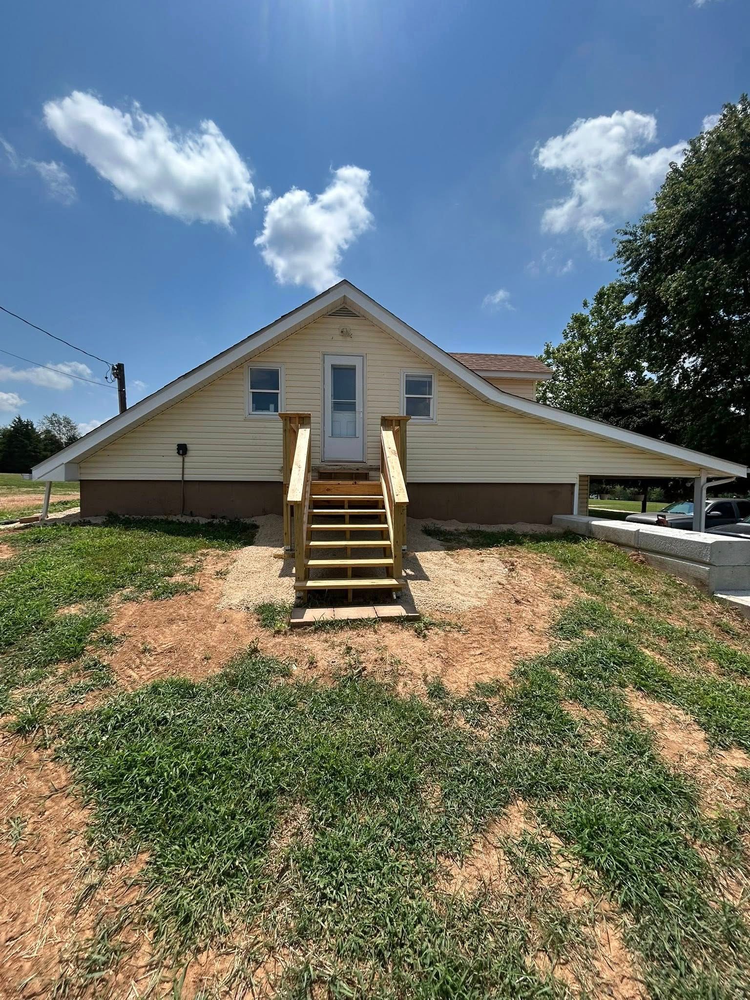 Tan house with wooden steps, carport, and blue sky.