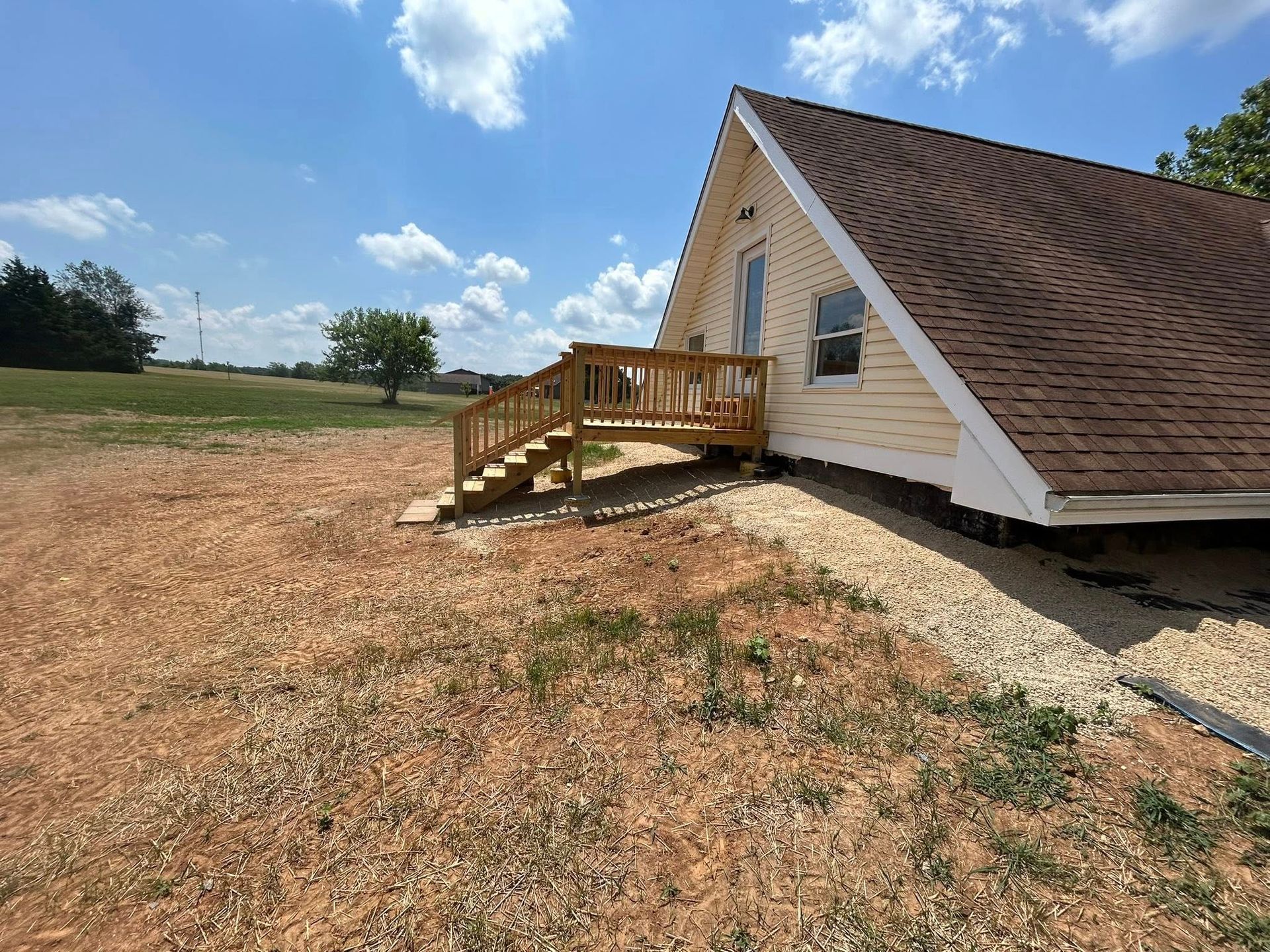 A-frame house with a wooden deck and steps; gravel landscaping in a sunny, rural setting.