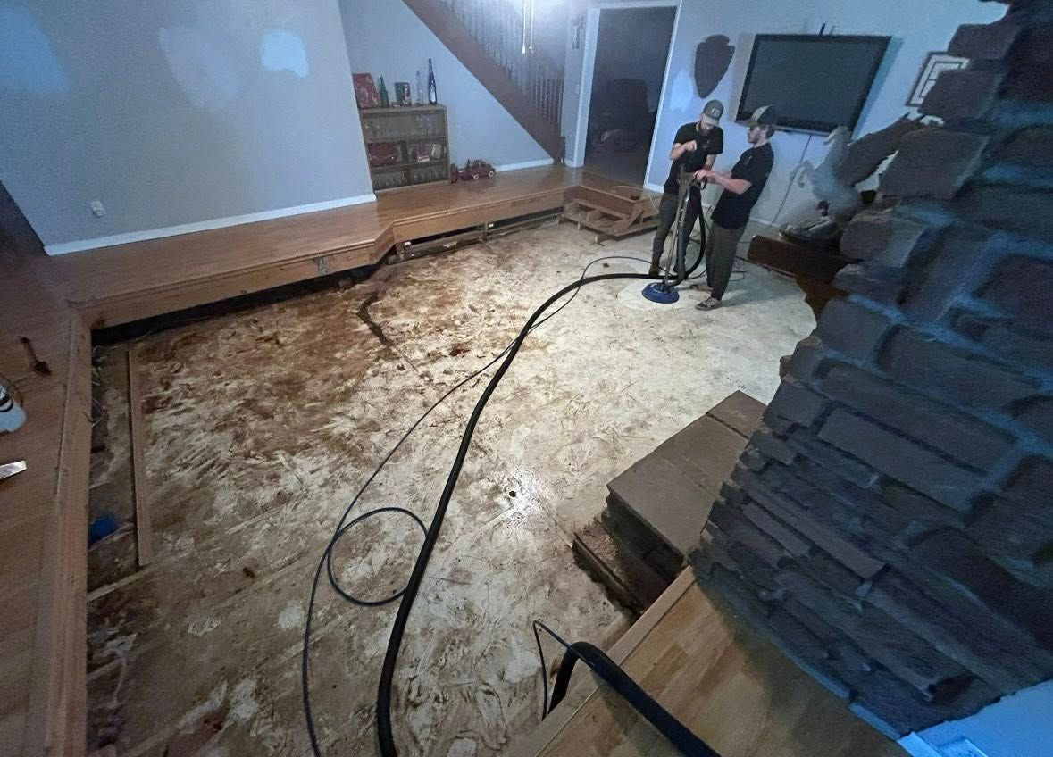 Two people vacuuming debris from a flooded room with damaged flooring. A dark hose stretches across the floor.