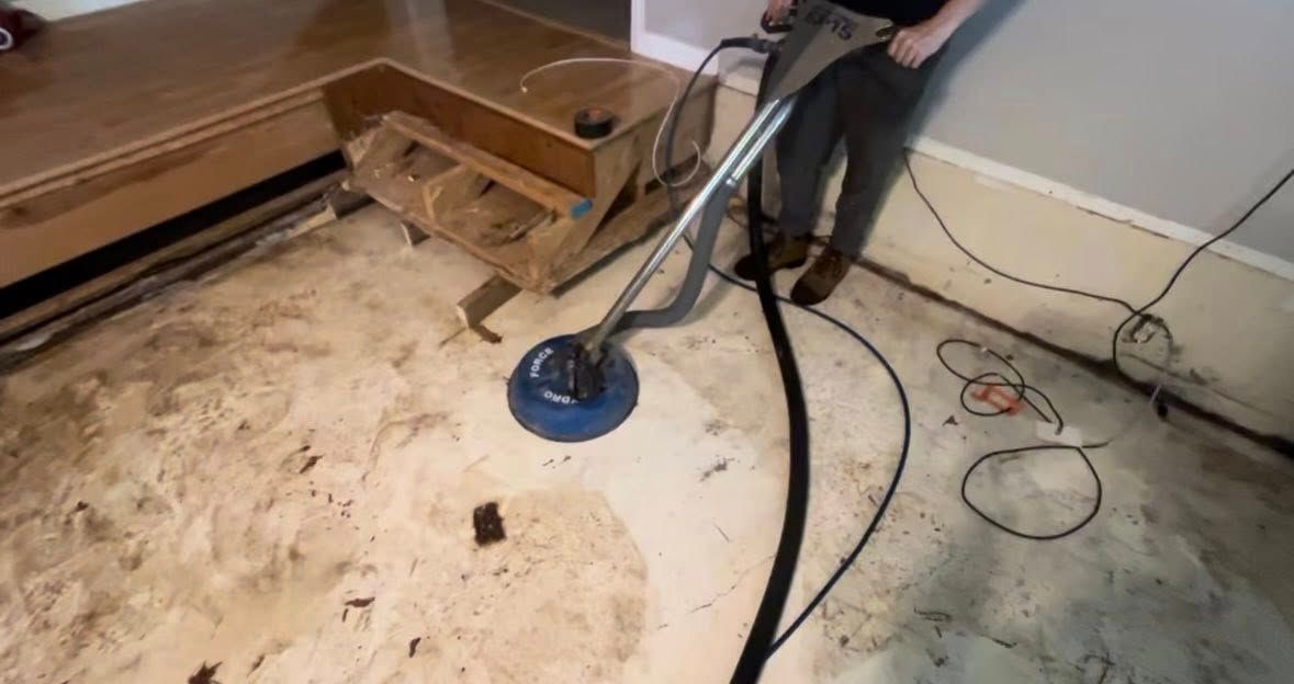 Person using floor cleaning machine on a concrete floor in a room under renovation.