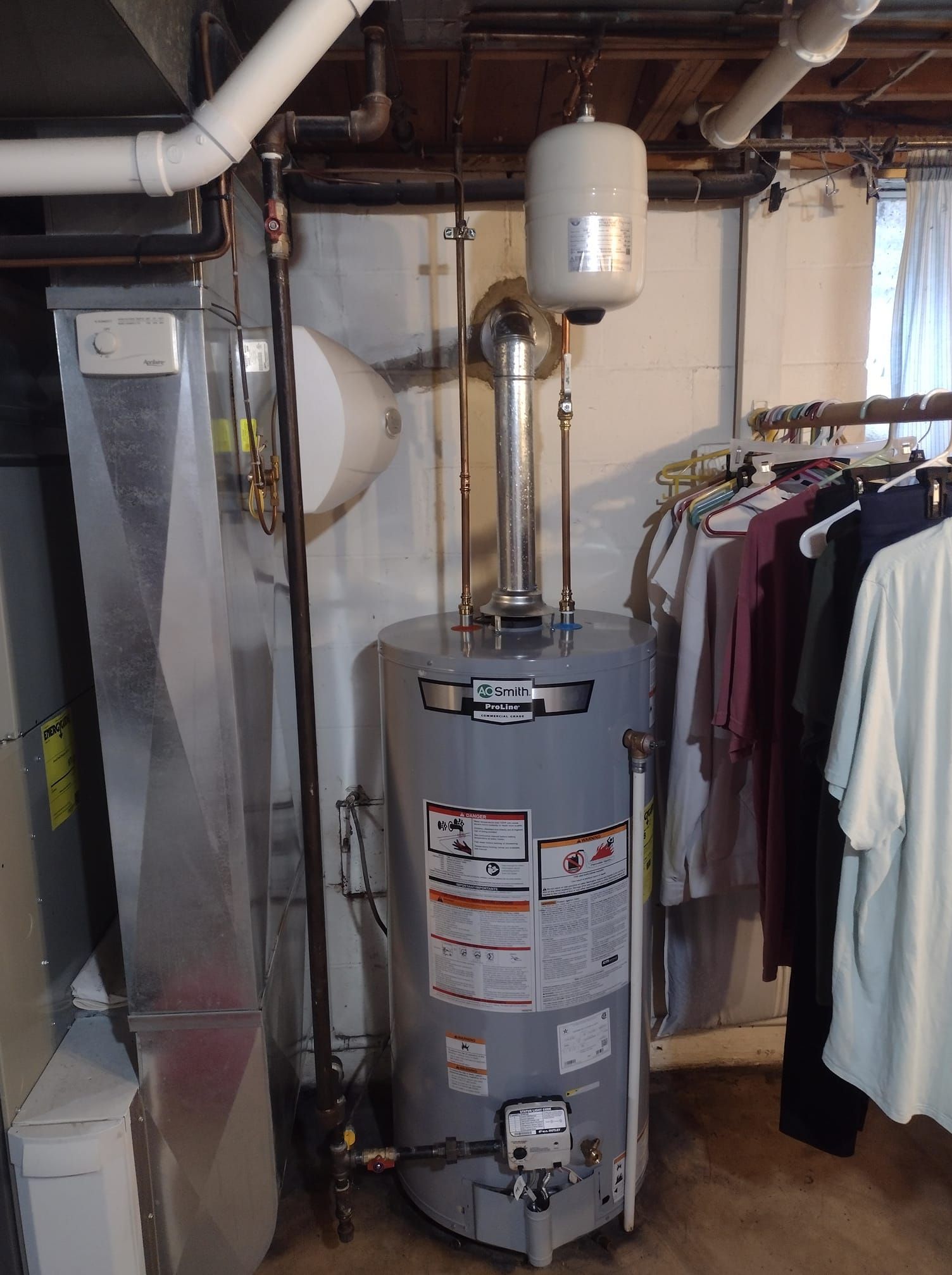 A water heater is sitting in a basement next to a clothes rack.