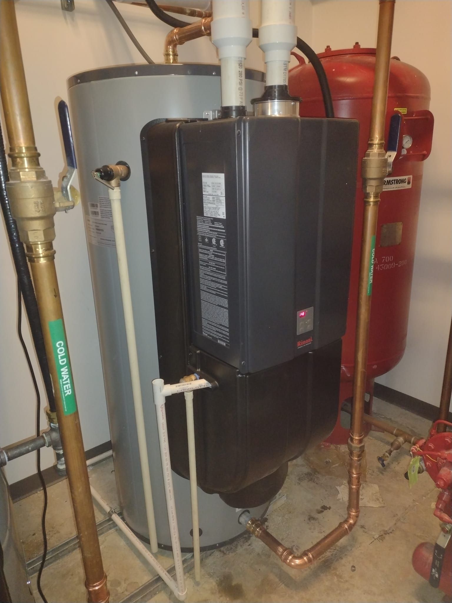 A water heater is sitting in a basement next to a red tank.