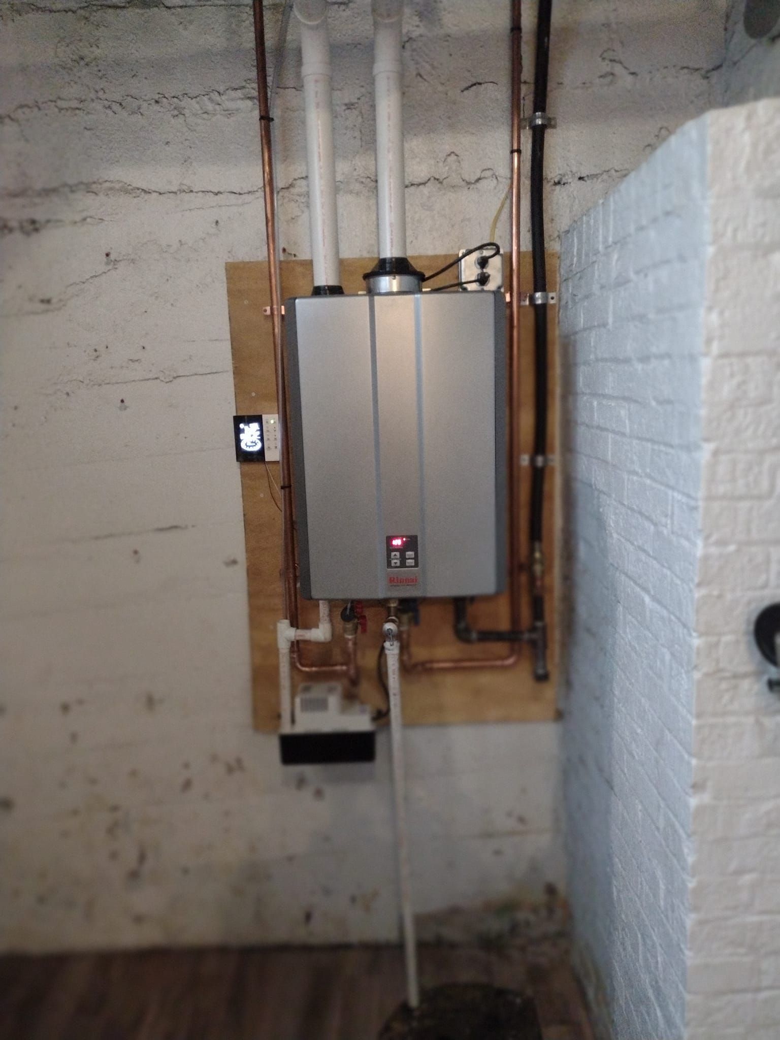 A water heater is installed in a basement next to a brick wall.