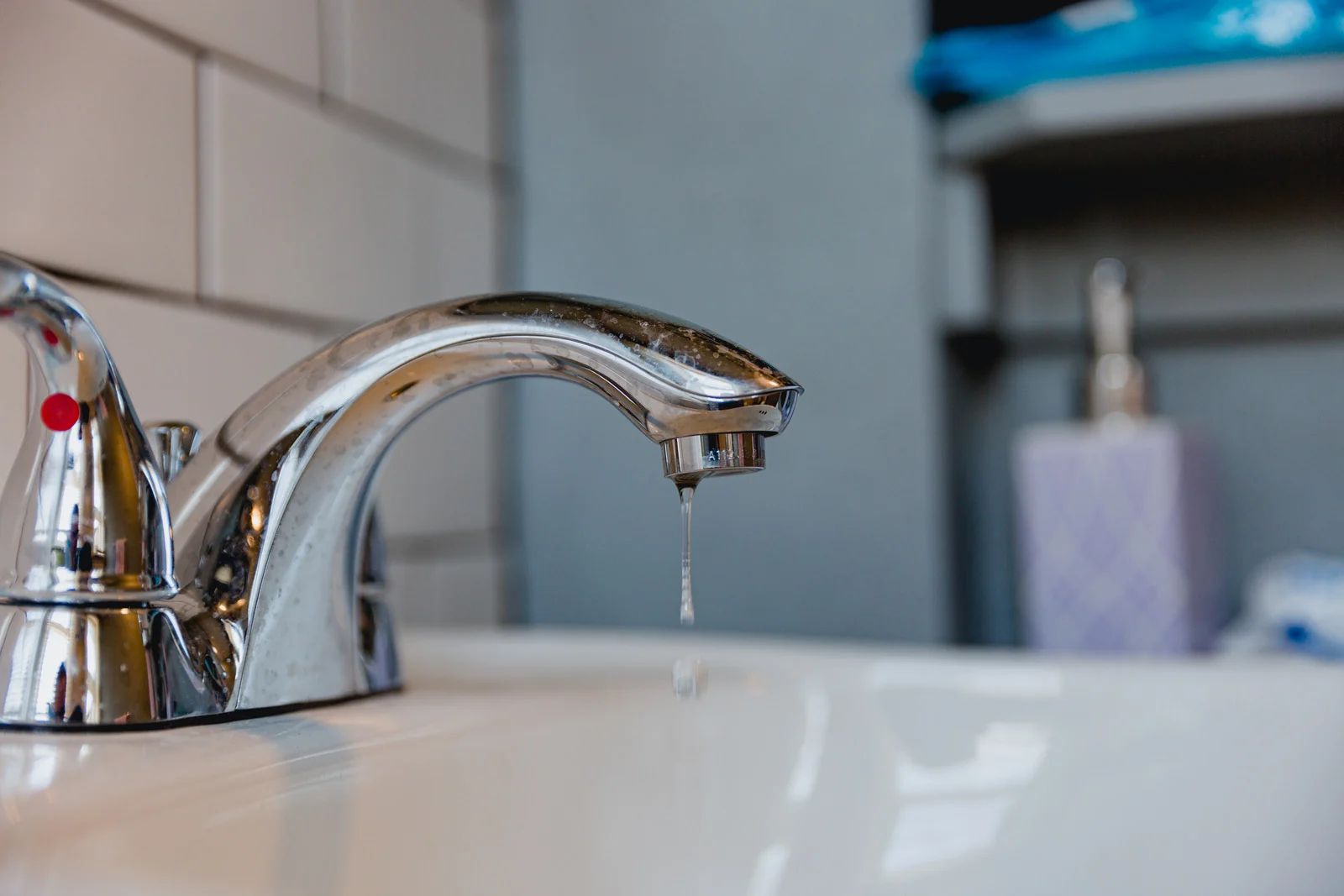 Chrome faucet with a water droplet hanging; set in a bathroom with a white sink.