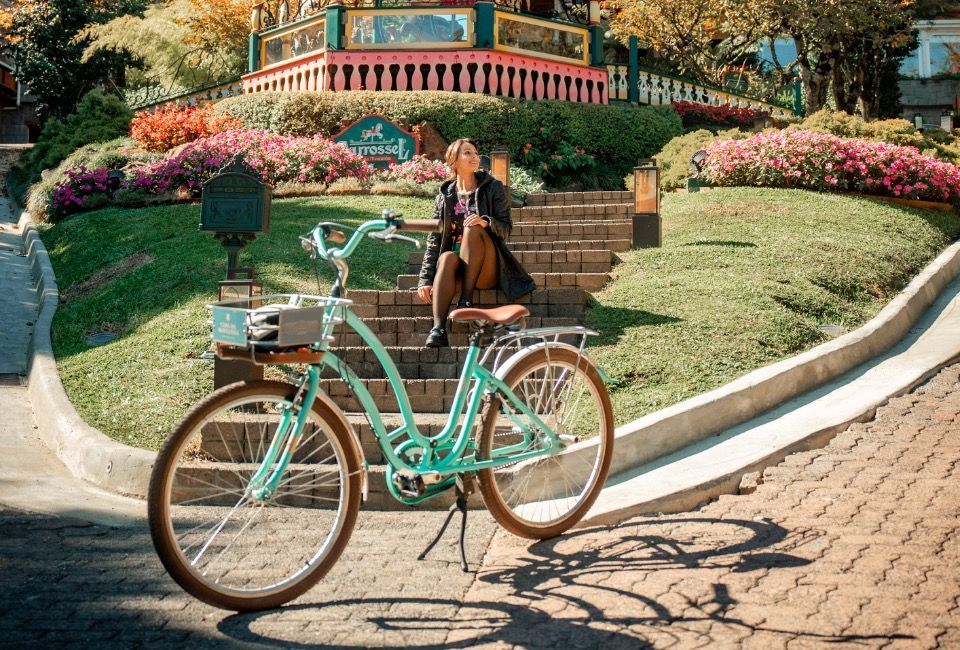Bicicleta verde-menta estacionada em um jardim ensolarado com uma pessoa sentada em degraus perto de flores cor-de-rosa.