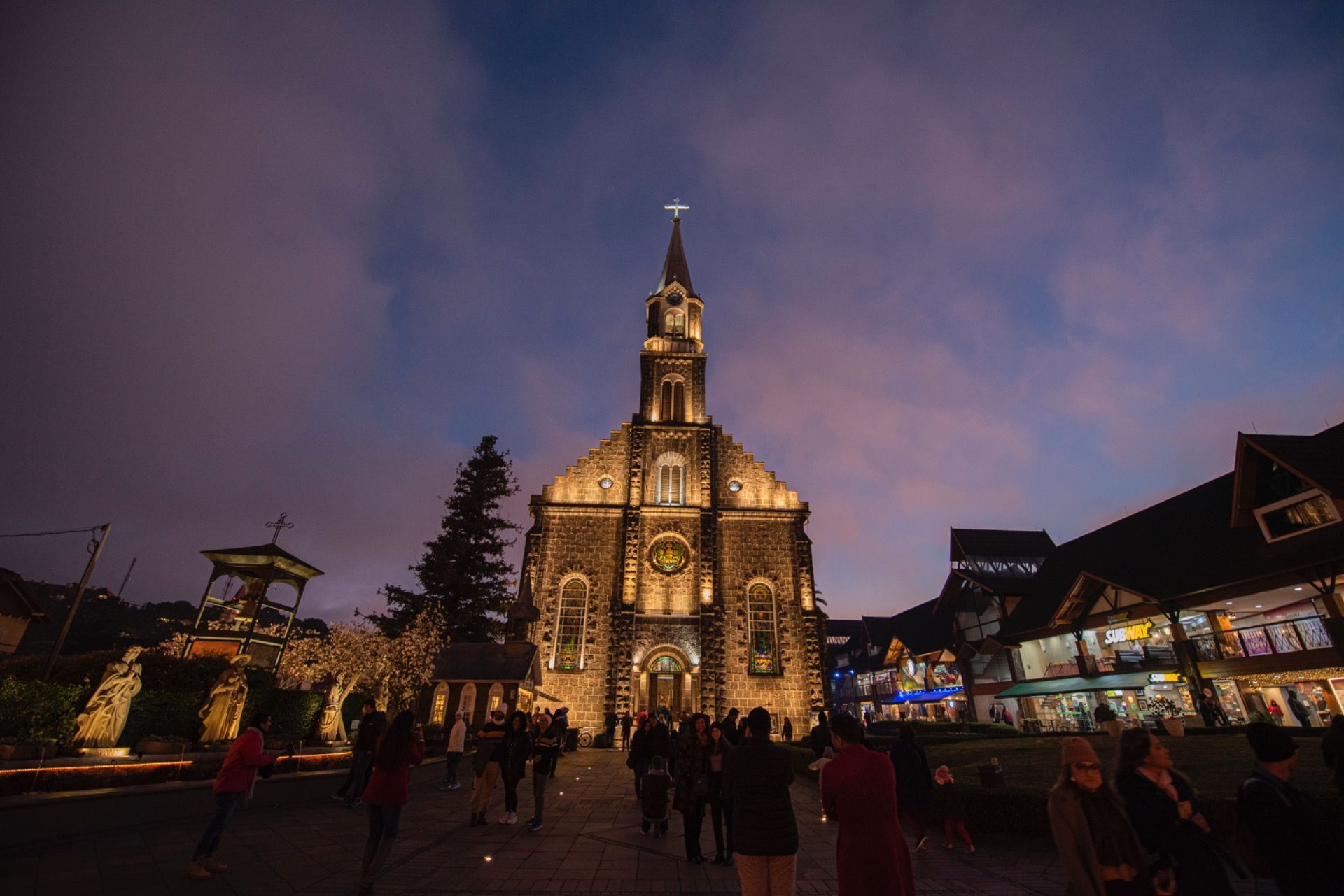 Igreja iluminada ao entardecer em uma praça festiva, com pessoas reunidas sob um céu púrpura.
