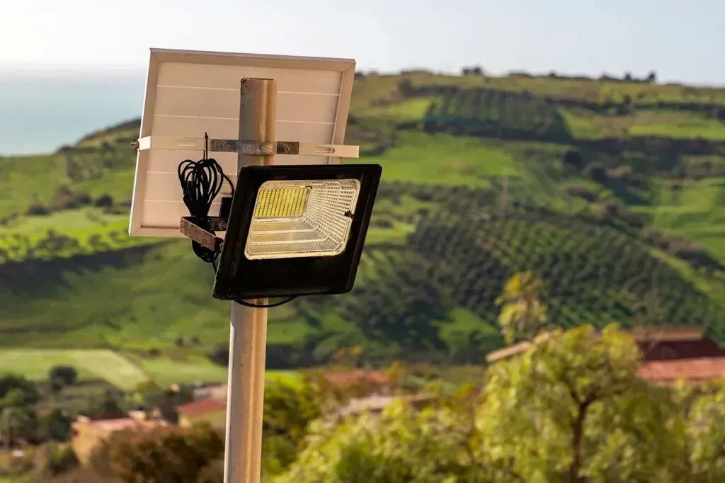 Un reflector LED alimentado por energía solar, montado sobre un poste, con un paisaje de colinas verdes y campos agrícolas de fondo.