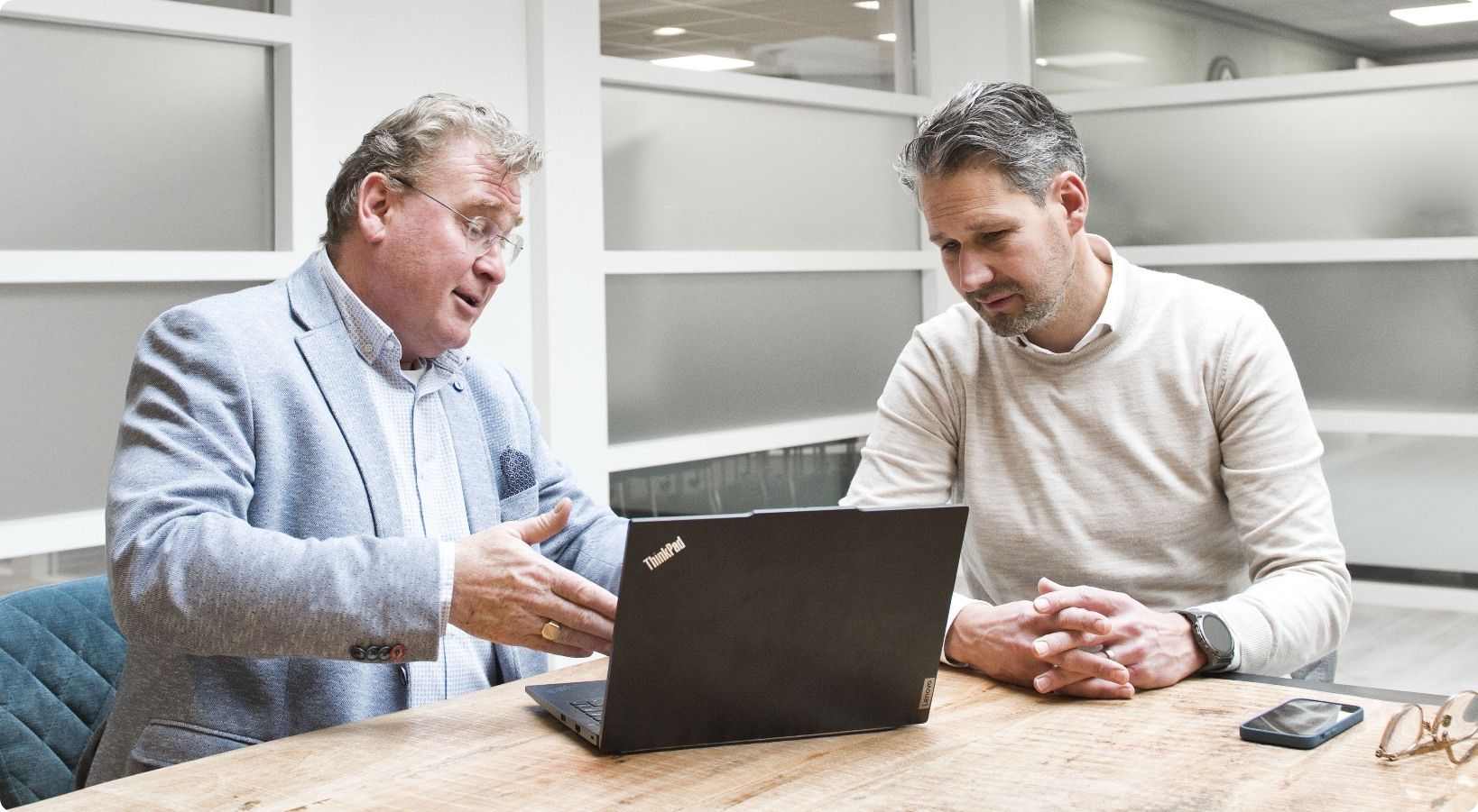 Two men at a table, looking at a laptop. The man on the left gestures, speaking. Neutral office setting.