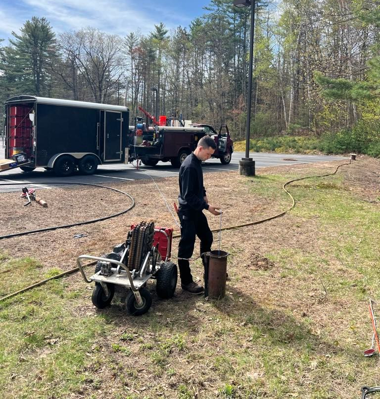 Man near equipment and a pipe, with a truck and trailer in the background on grass.
