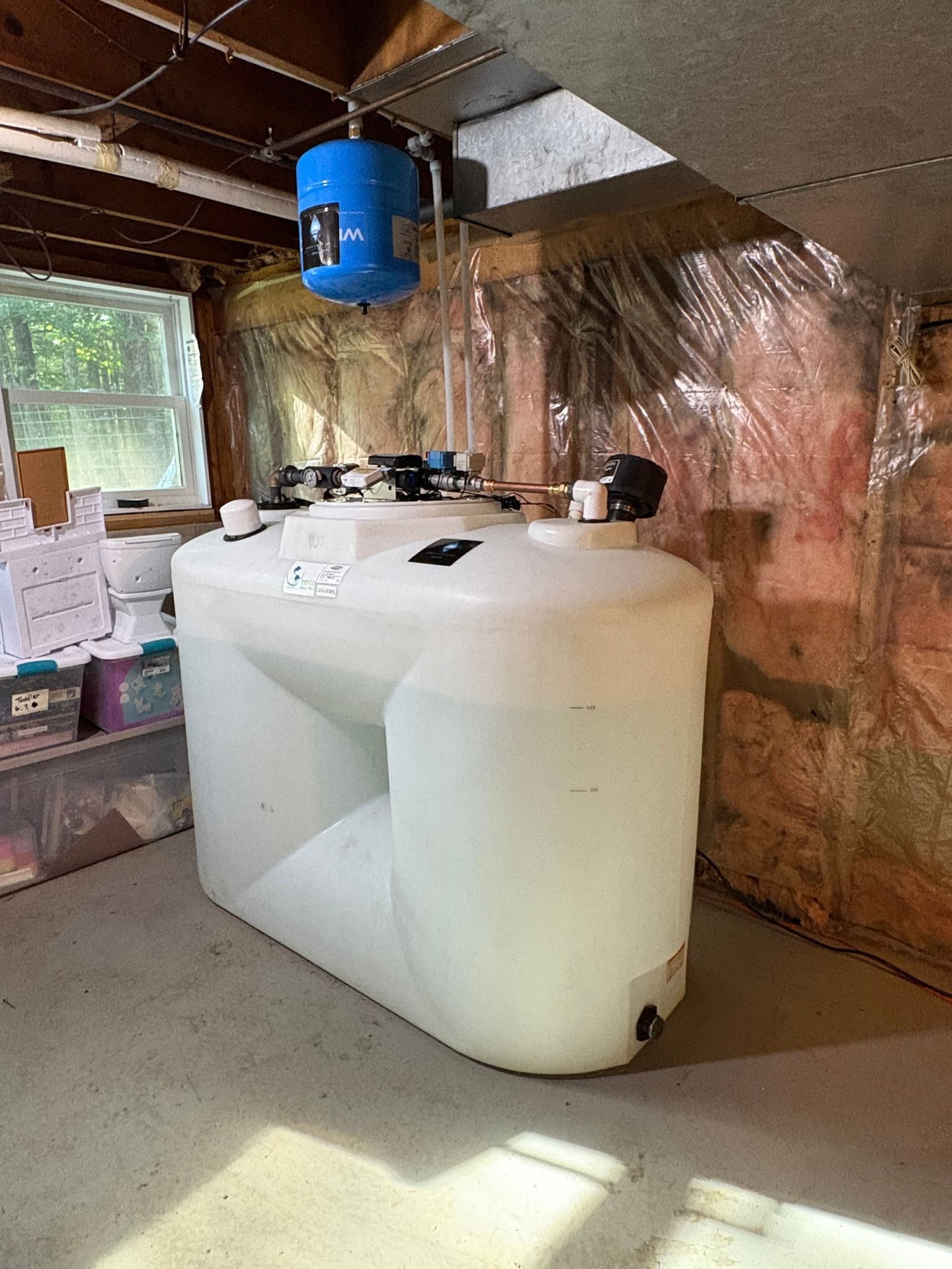 Large white water tank in a basement with attached blue pressure tank and piping.