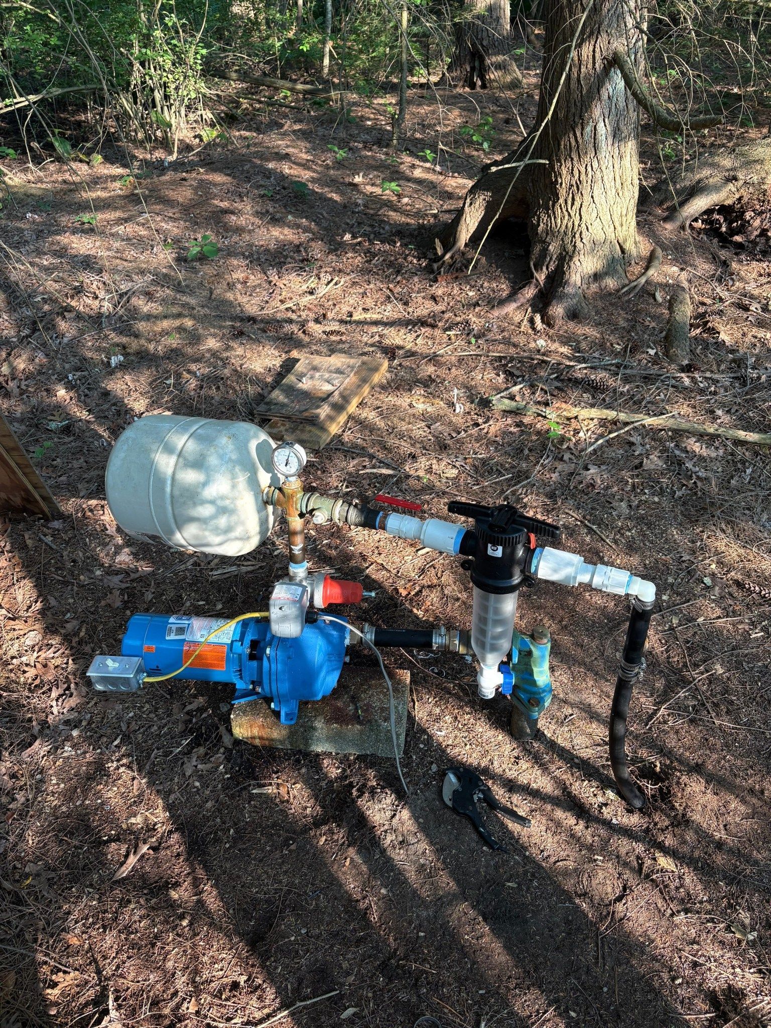 Well pump system outdoors, with blue pump, white tank, and filter on brown ground near trees.